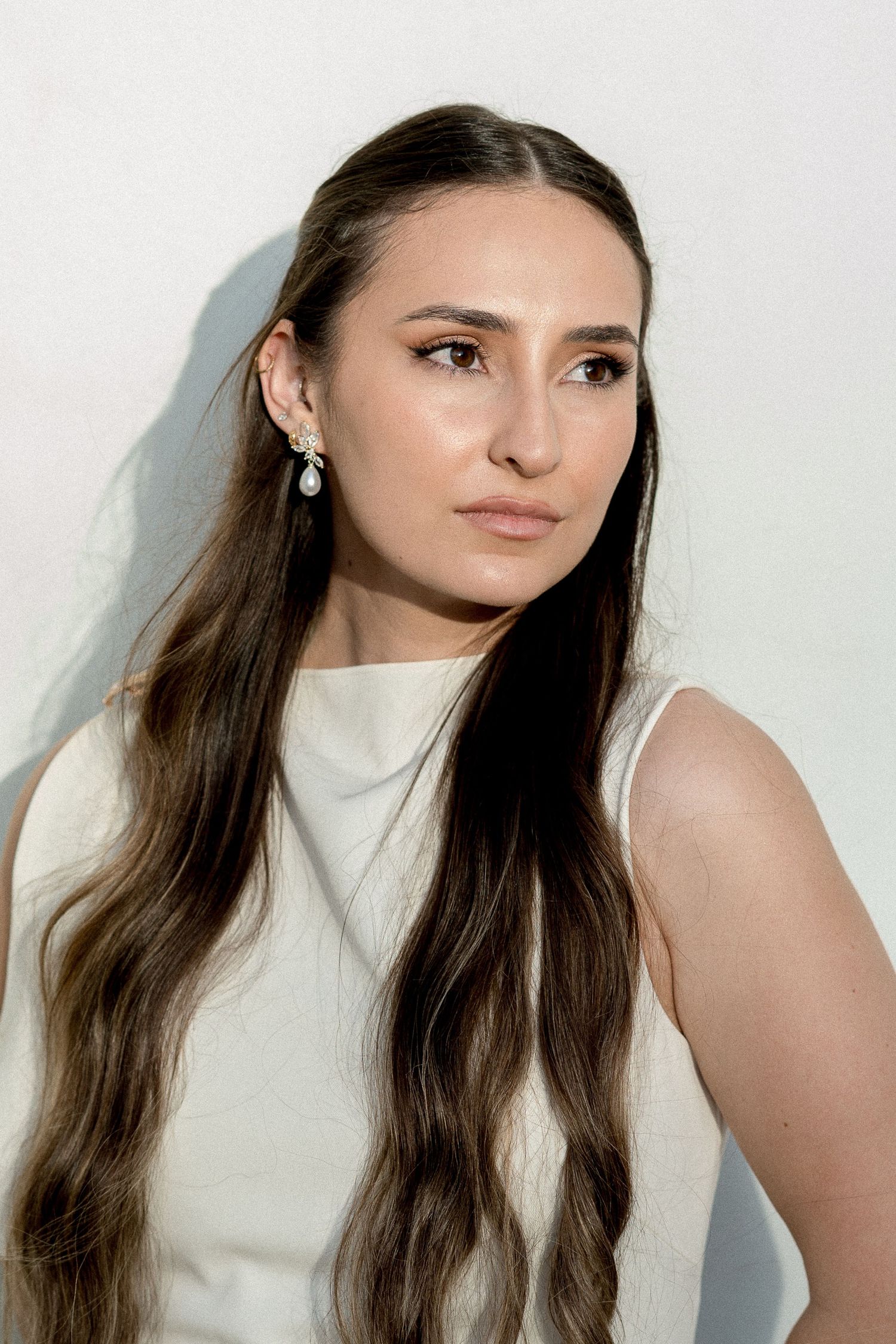 Young woman with long wavy brown hair wearing a white sleeveless top and drop earrings against a light background.