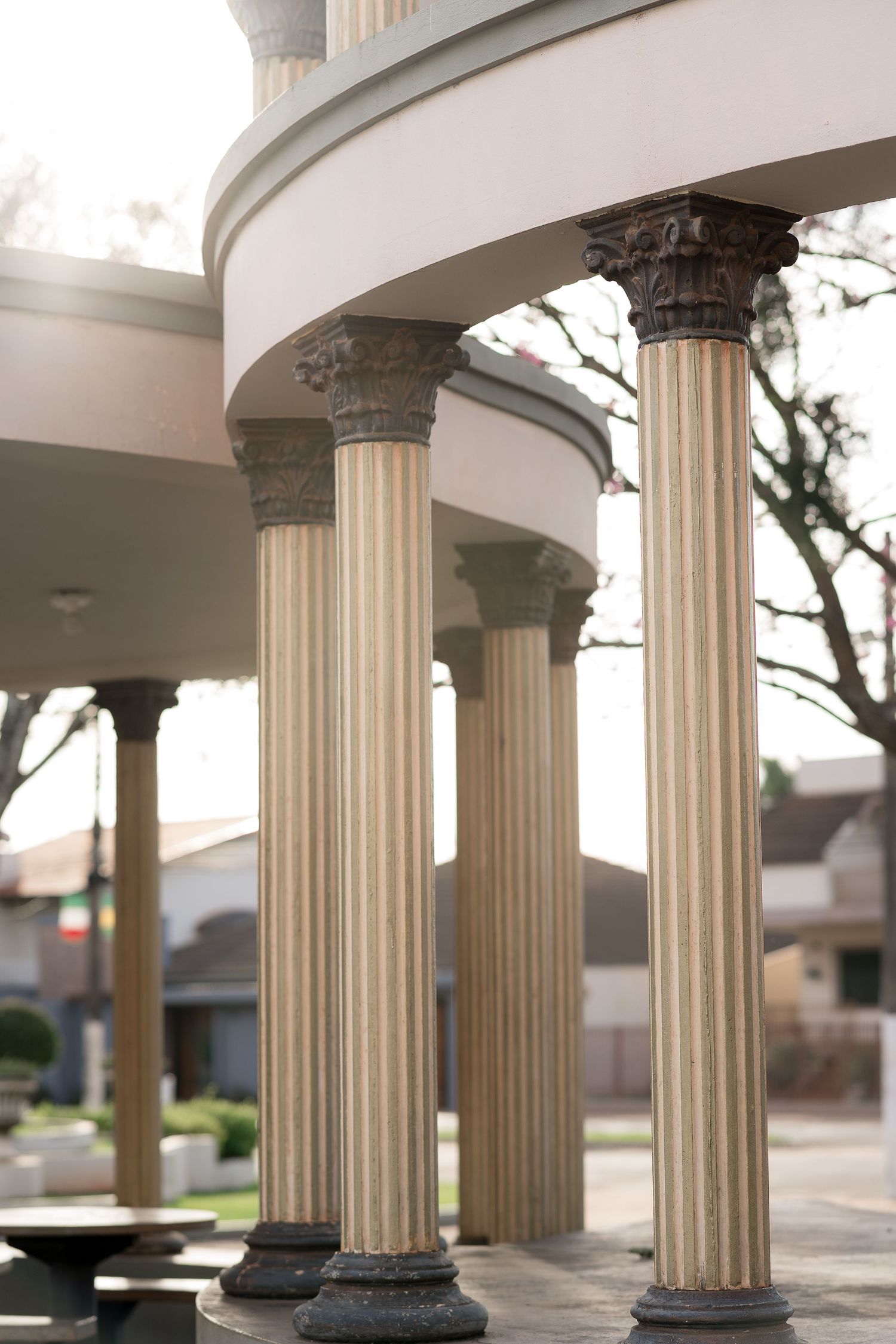 Classical fluted columns with ornate Corinthian capitals support a curved rotunda structure in an outdoor park setting.