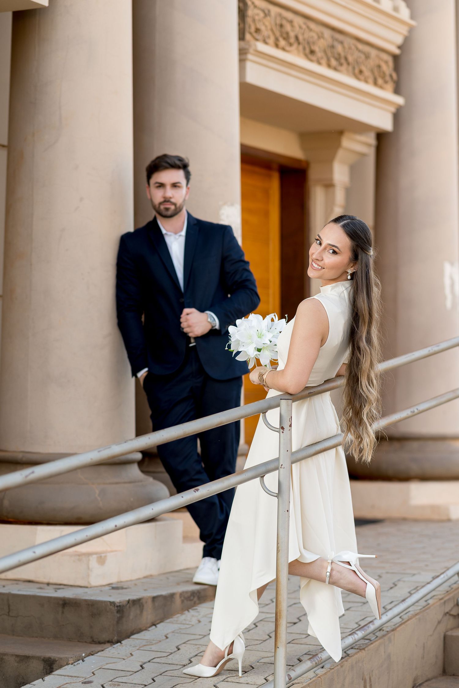 Elegant bride in white dress holding bouquet poses on steps with groom in dark suit near classical columns.