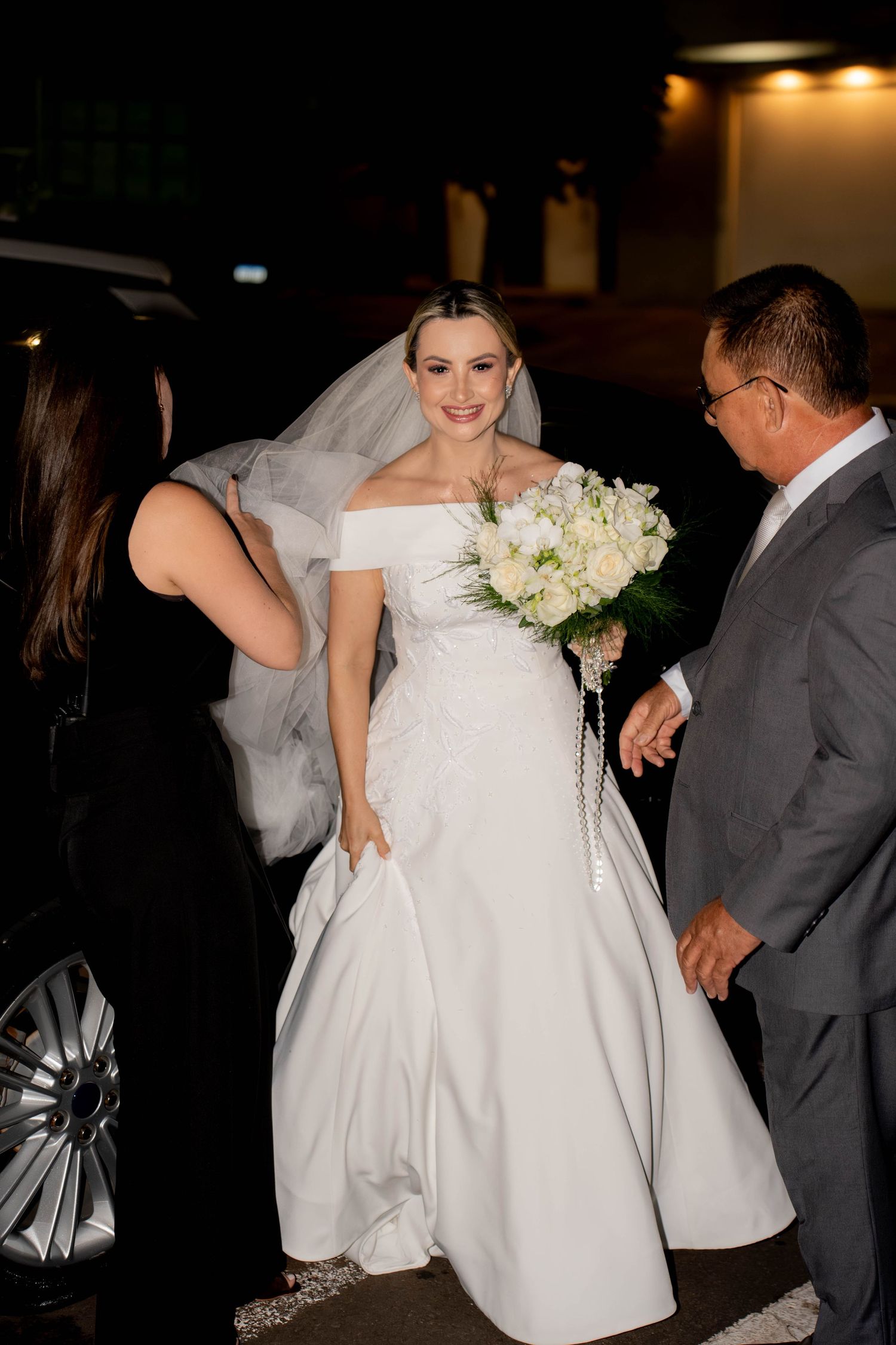 Smiling bride in white off-shoulder gown and veil holds bouquet of white flowers while being assisted by a man in gray suit.