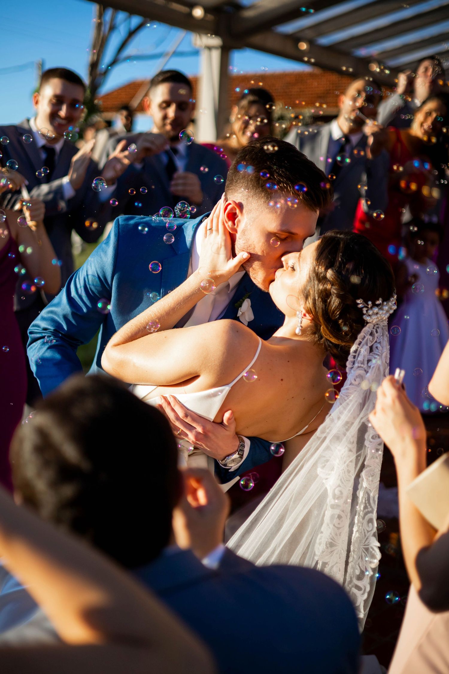 Newlyweds share a romantic kiss surrounded by colorful soap bubbles and cheering wedding guests outdoors.