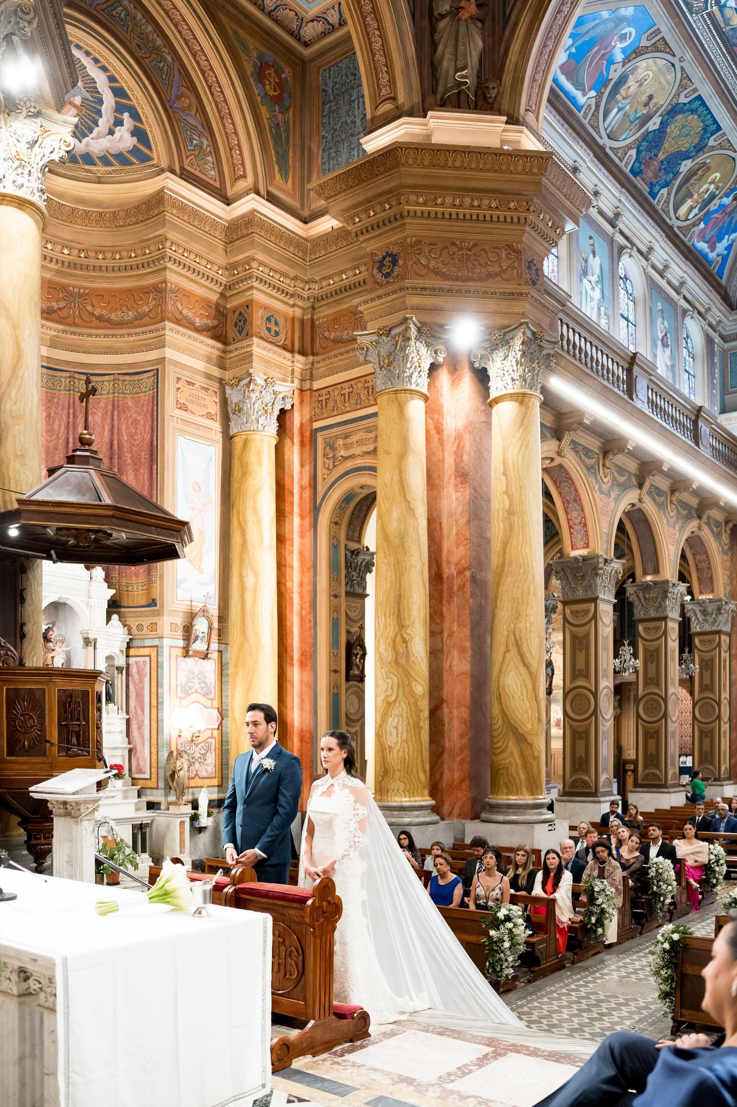 Bride and groom stand at the altar of an ornate Italian church with marble columns and frescoed ceilings.