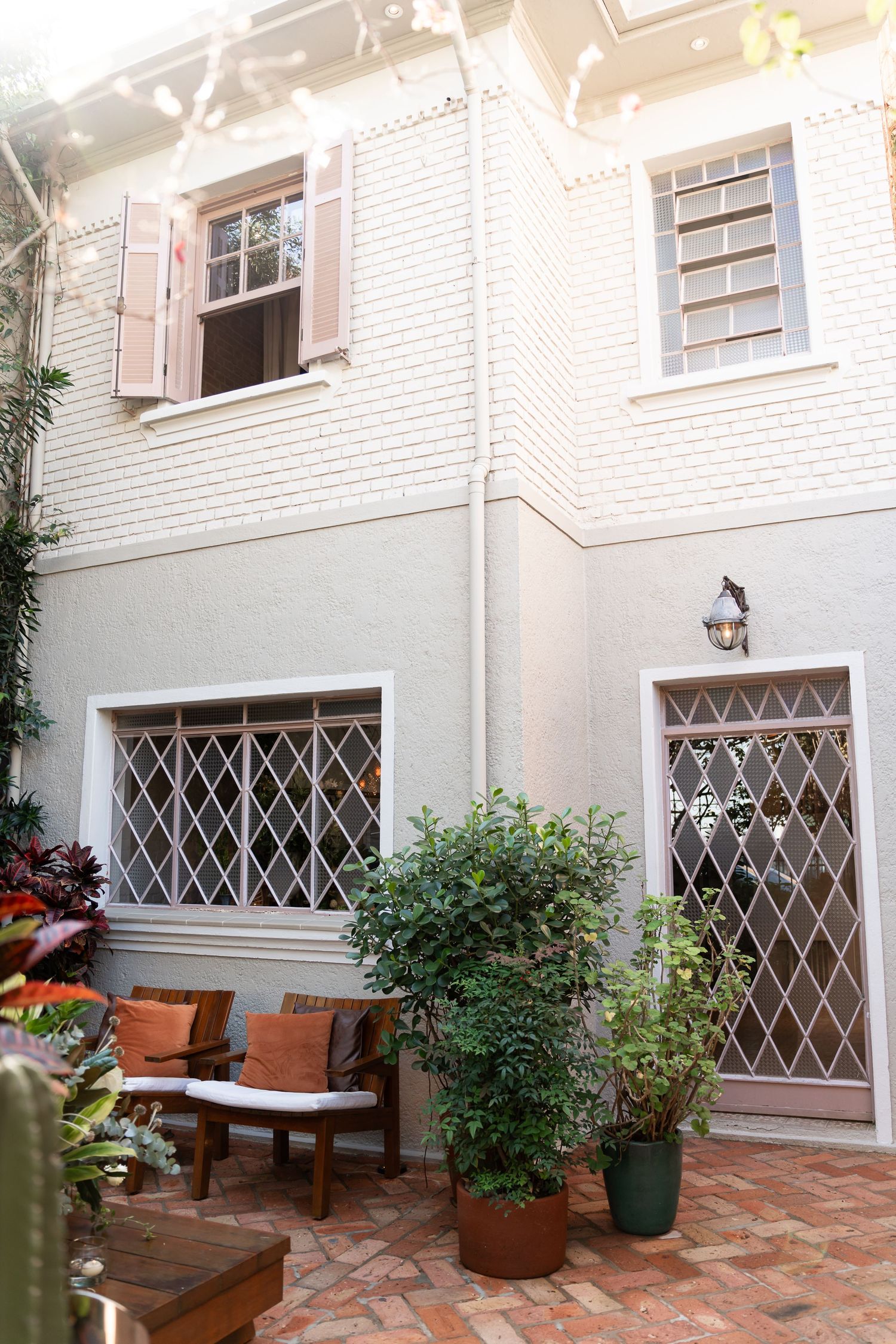 Charming courtyard patio with brick flooring, diamond-grill windows, potted plants, and a wooden bench with orange cushions.