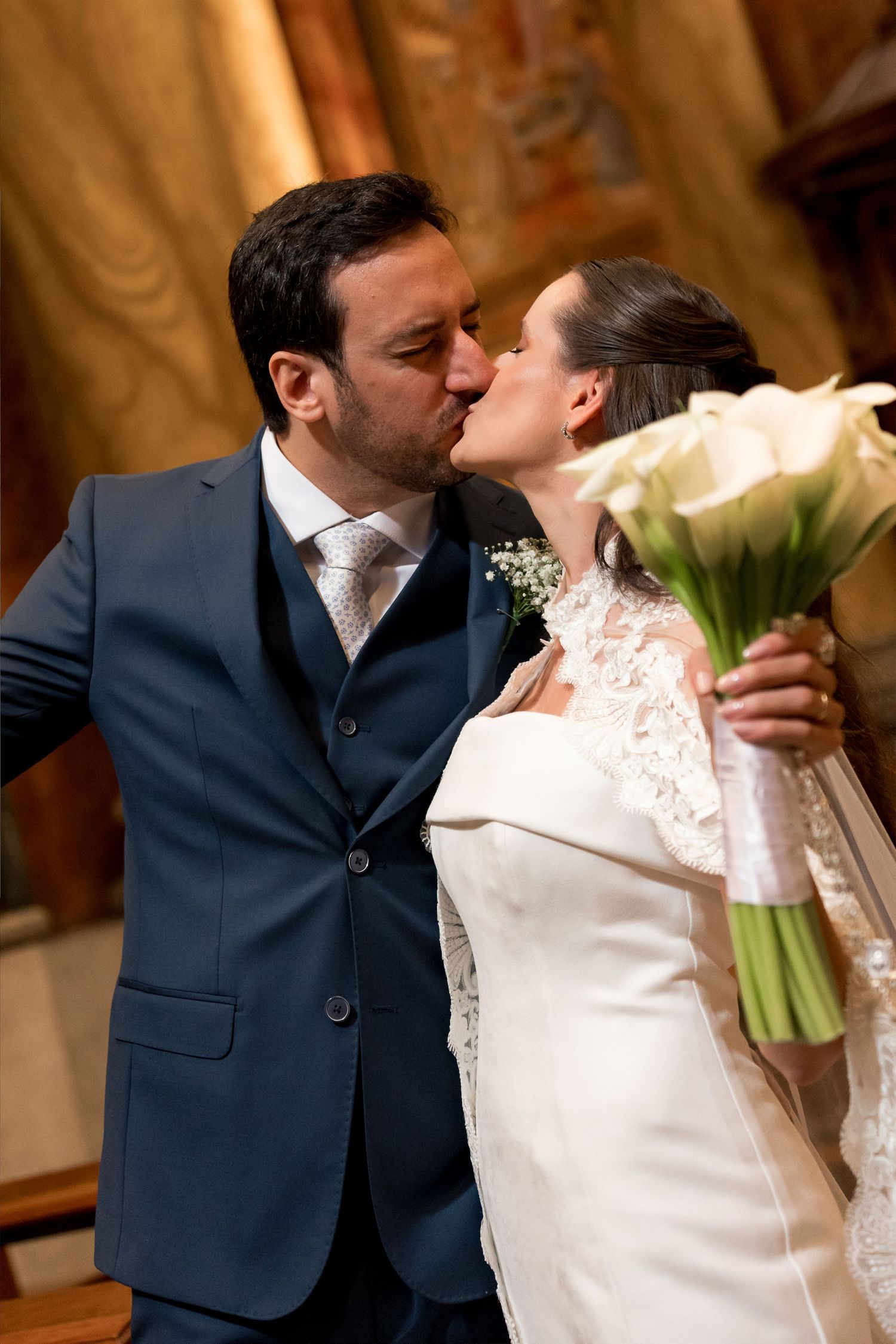 Bride and groom share a kiss at their wedding, bride holding white calla lily bouquet, groom in navy suit.