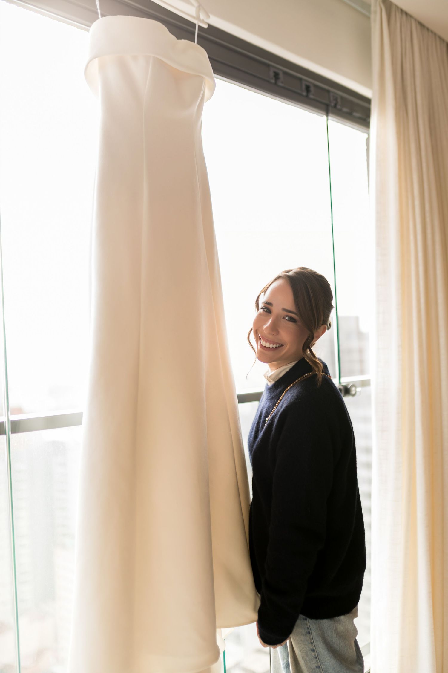 Smiling bride-to-be in navy sweater standing beside her elegant white wedding gown hanging by a bright window.
