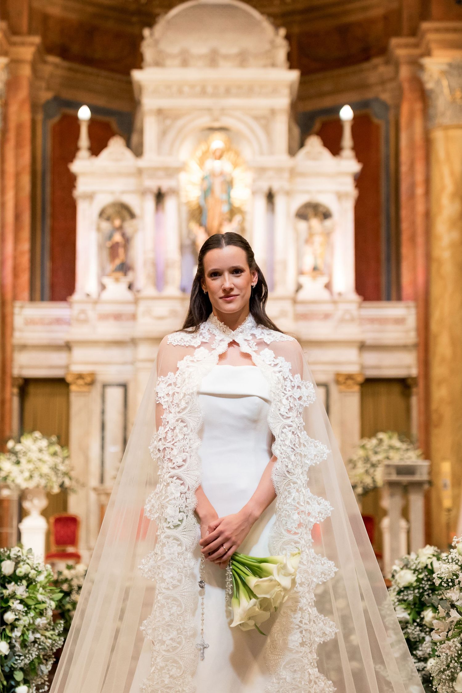Bride in white gown with lace cape holds bouquet before ornate church altar adorned with floral arrangements.