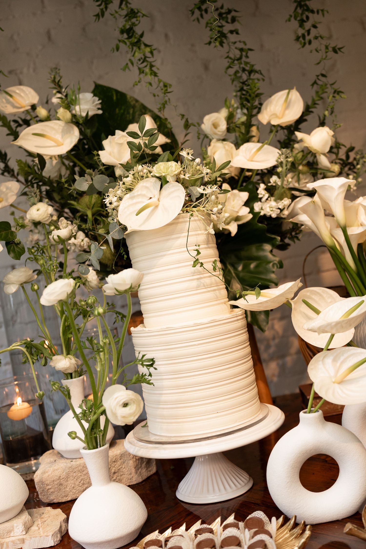 Elegant three-tier white wedding cake surrounded by white calla lilies and ranunculus floral arrangements on a decorated table.