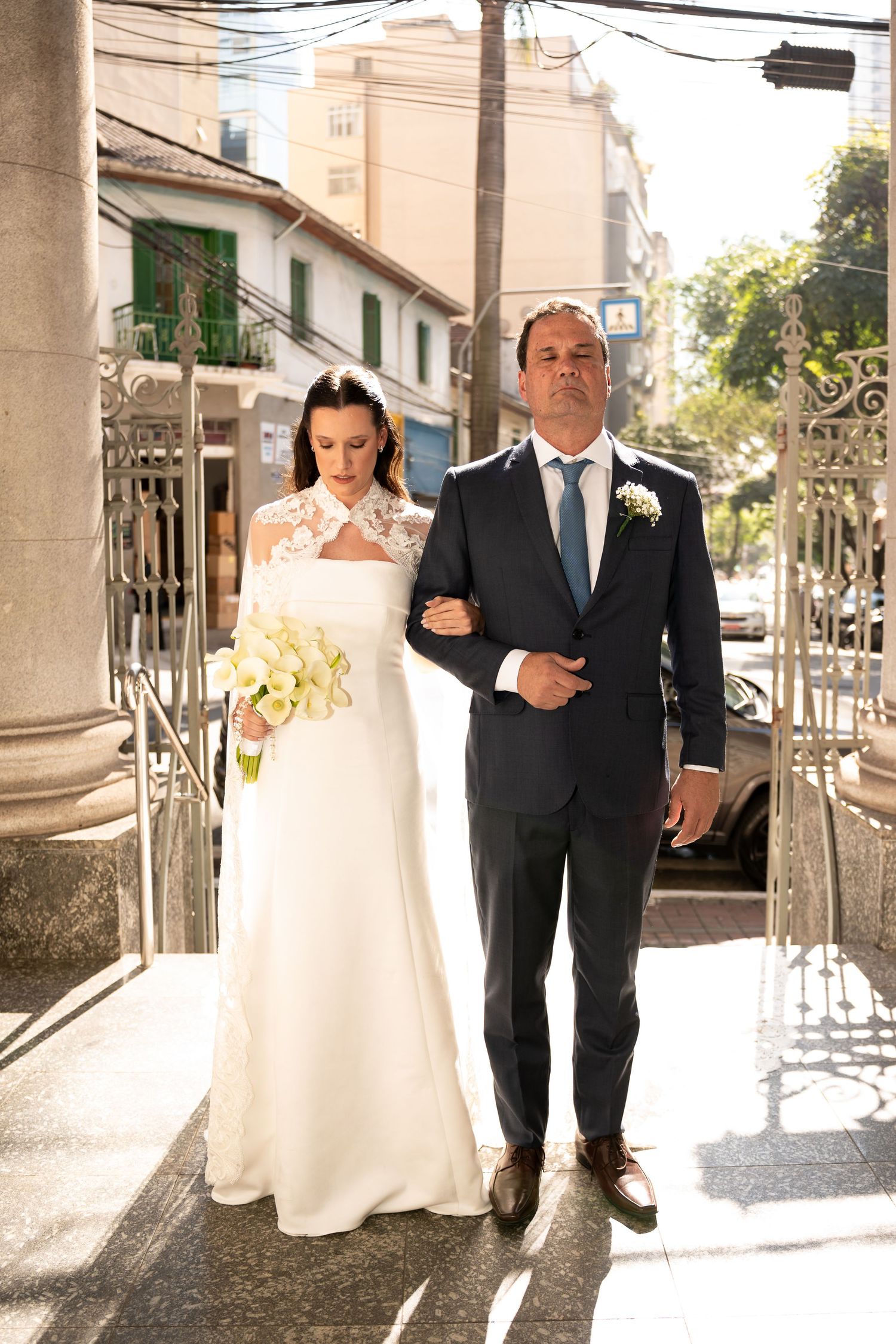 Bride in white gown holding yellow flowers walks with groom in dark suit outside a sunlit church entrance.