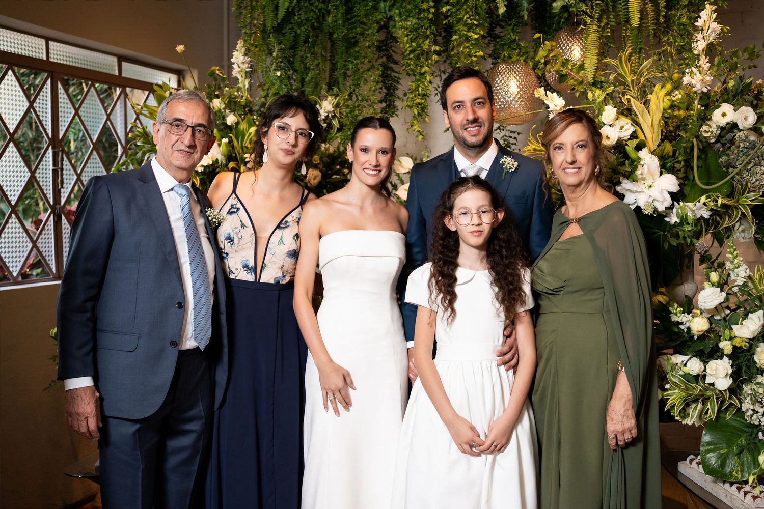 Wedding party posing with bride and groom surrounded by lush floral greenery backdrop at elegant indoor reception.