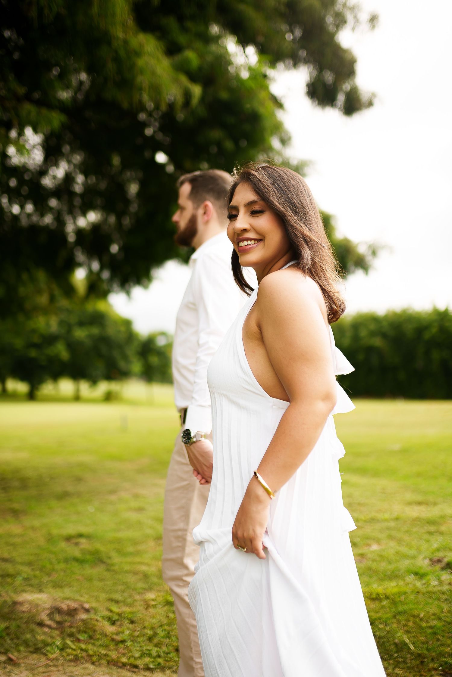 A smiling woman in a white dress stands in a park with a man in white shirt, lush green trees in background.