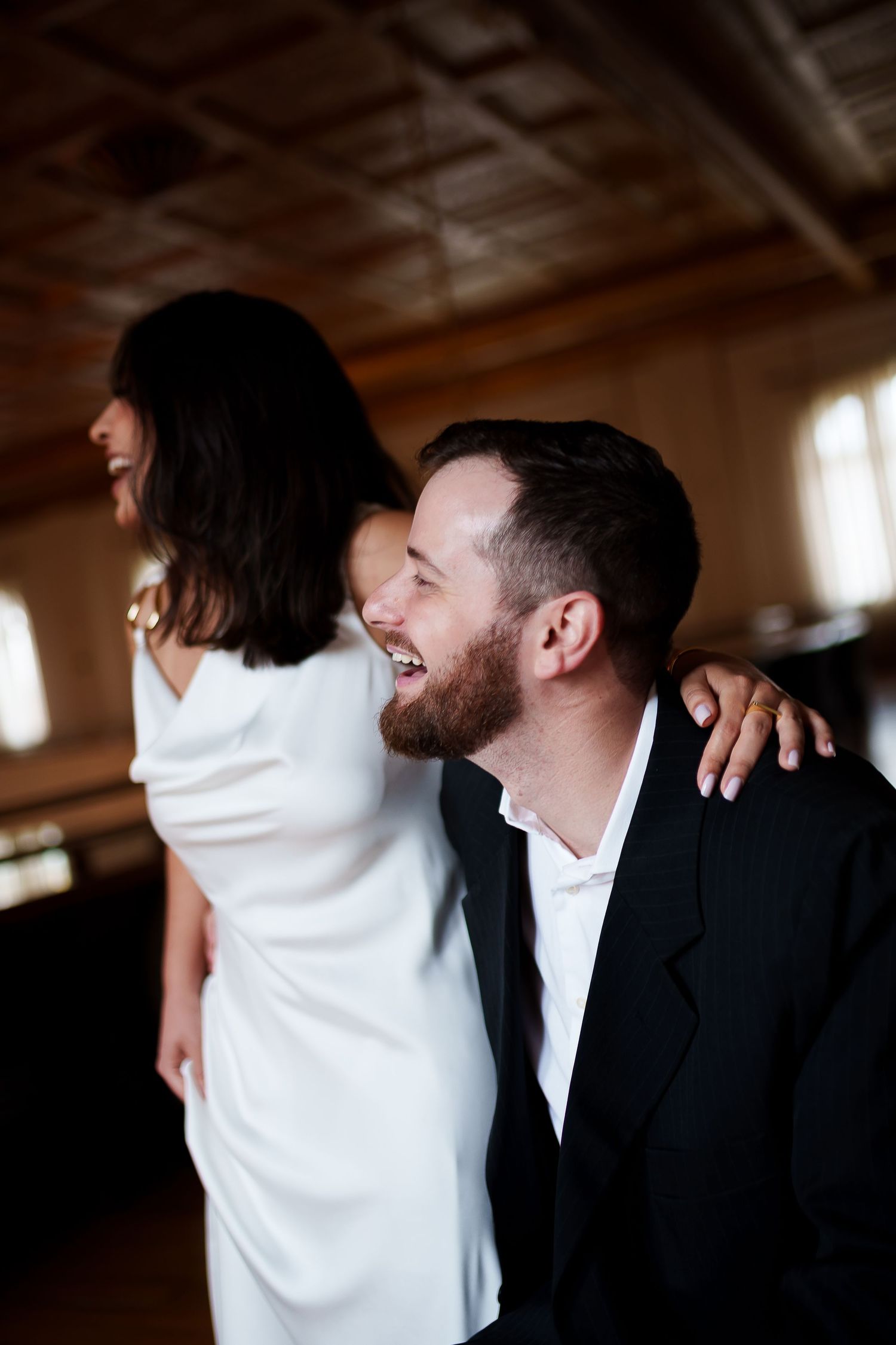 A laughing groom in a black suit embraces a bride in a white dress during their wedding celebration indoors.