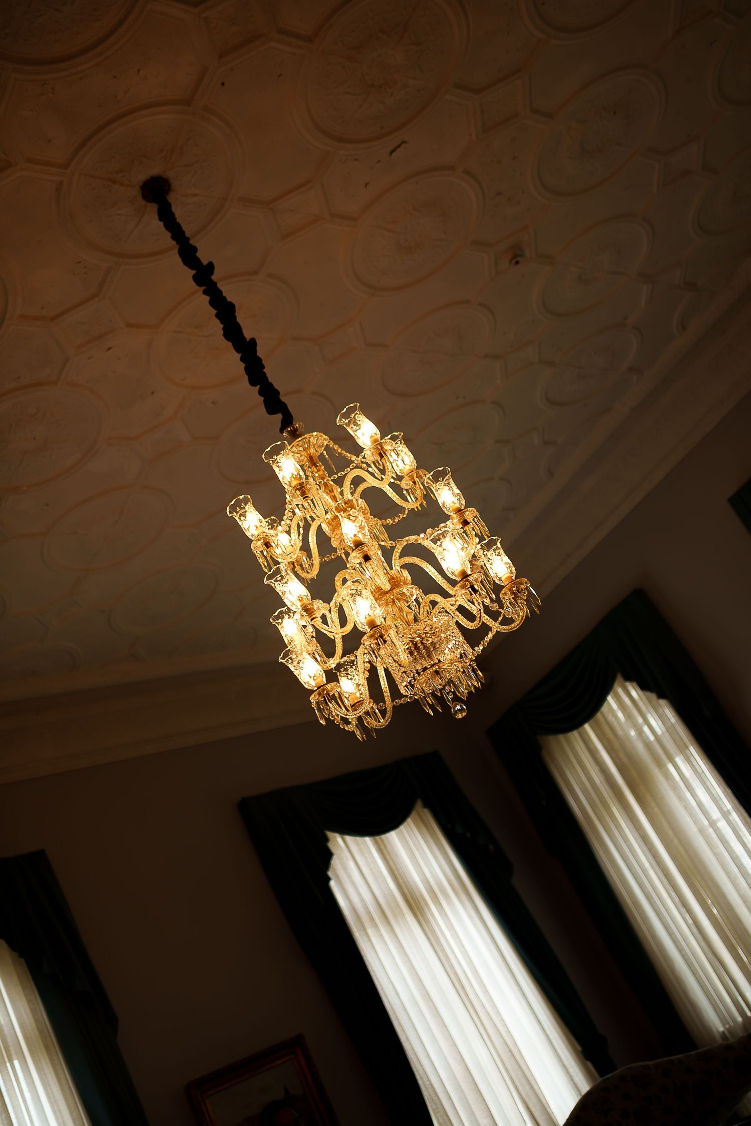 Ornate gold crystal chandelier with glowing candle-style lights hanging from decorative white plaster ceiling in elegant room.