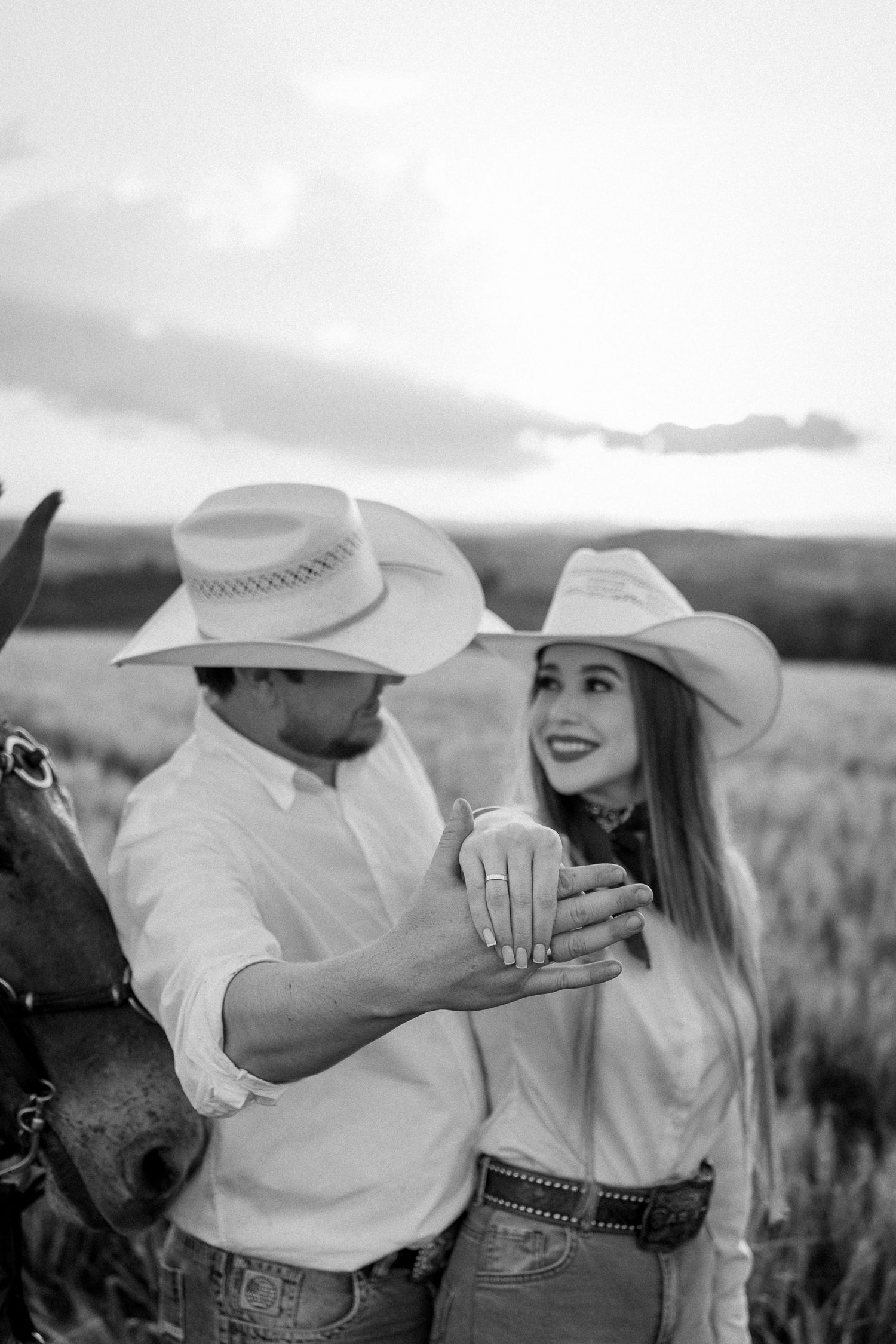 Black and white photo of a couple in cowboy hats examining an engagement ring in an open field.