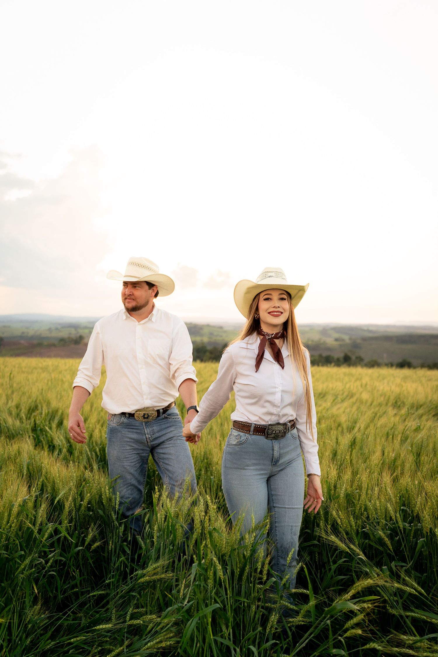 A couple in matching white shirts, jeans, and cowboy hats hold hands while walking through a lush green wheat field at sunset.