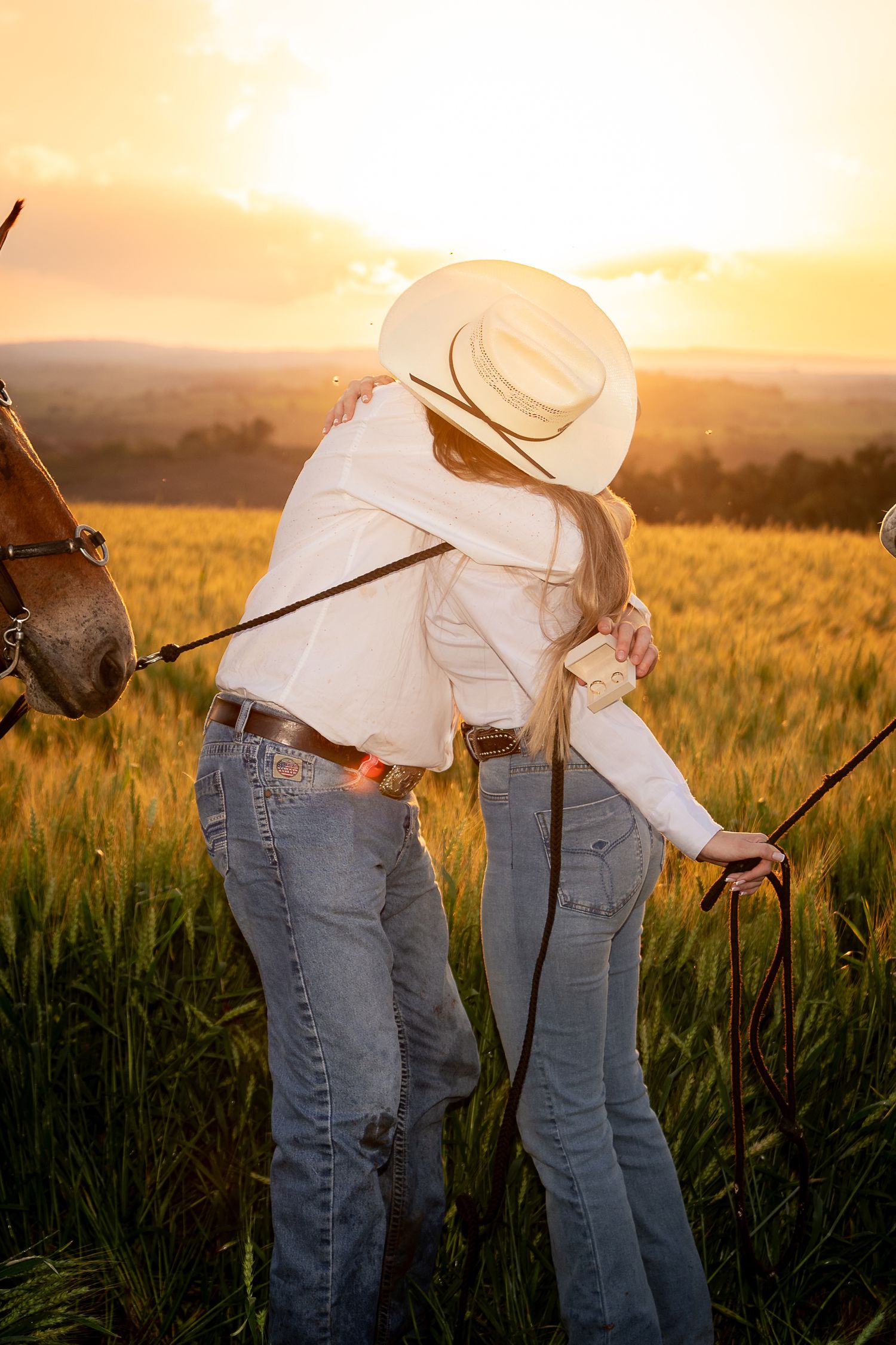 Two cowboys embrace in a golden wheat field at sunset, with a horse nearby, wearing white shirts and jeans.