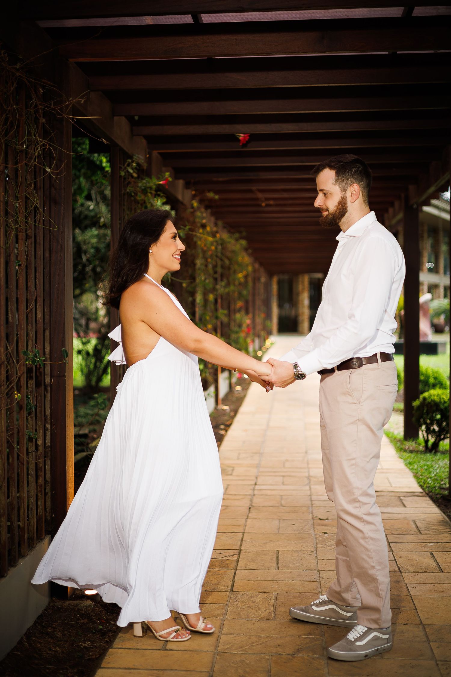 Couple in white dress and khakis holding hands under a wooden pergola on a stone pathway surrounded by greenery.