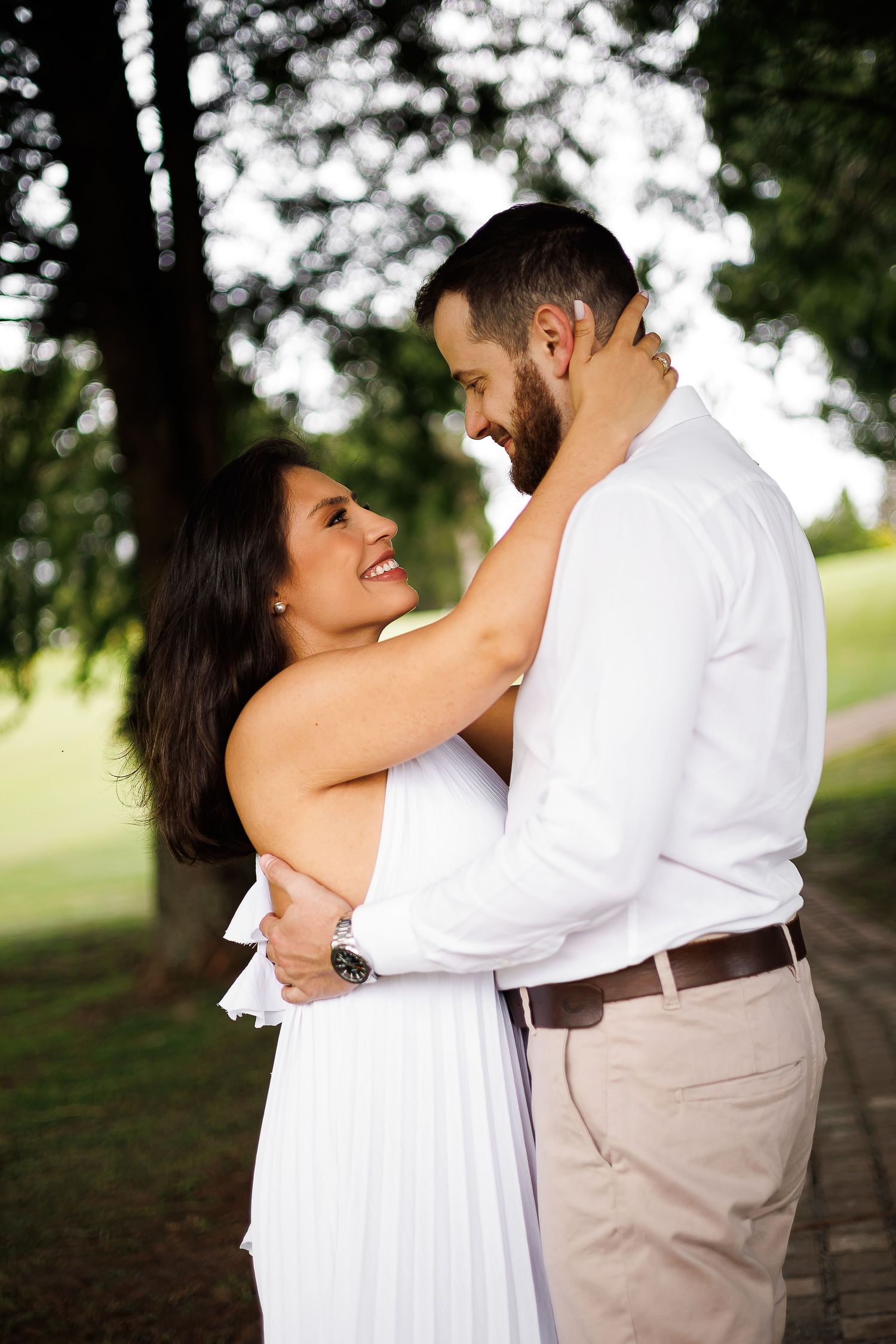 A couple embraces outdoors, smiling at each other, wearing white outfits, surrounded by lush green trees.
