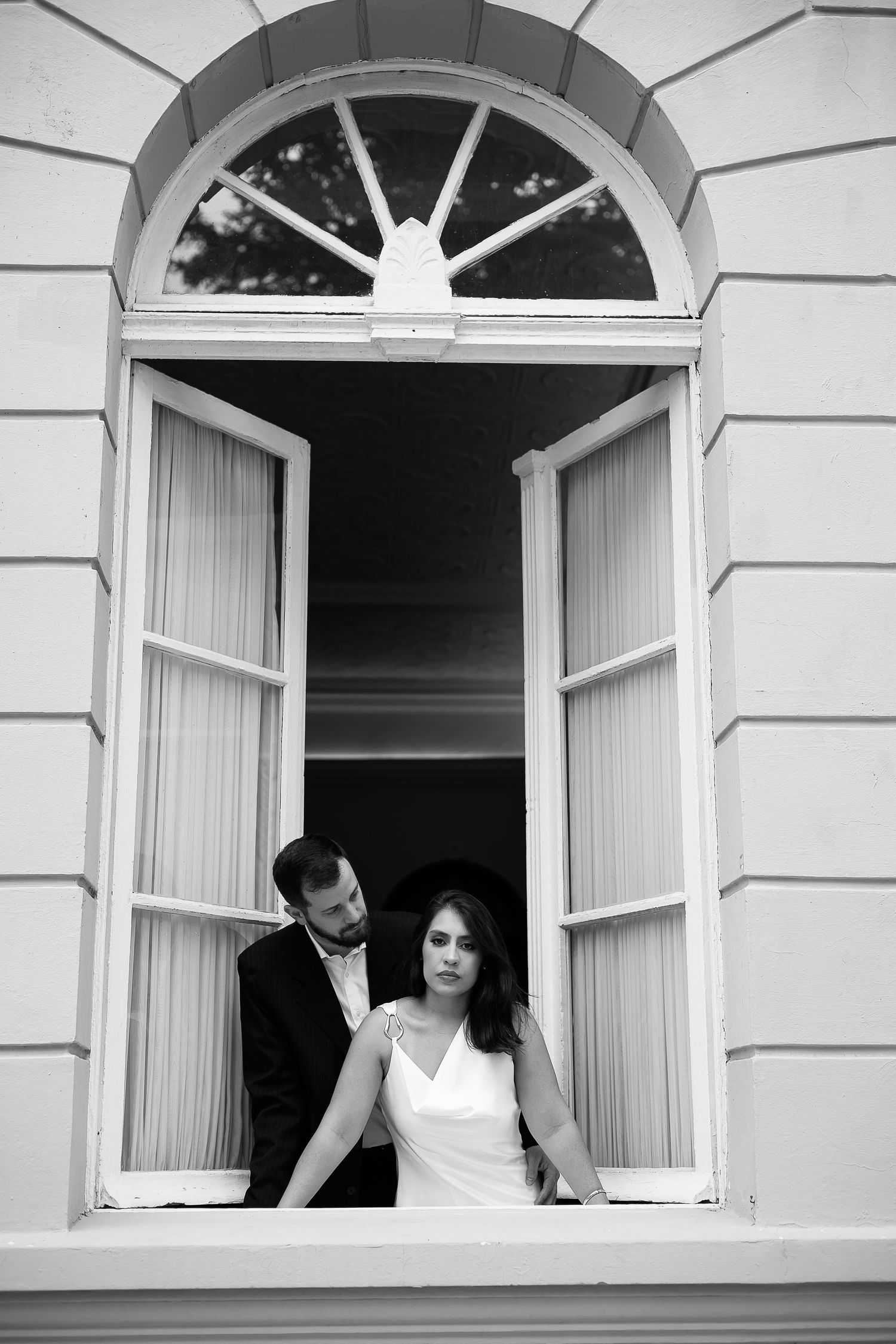 Black and white photo of a bride and groom posing in an open arched window of an elegant building.