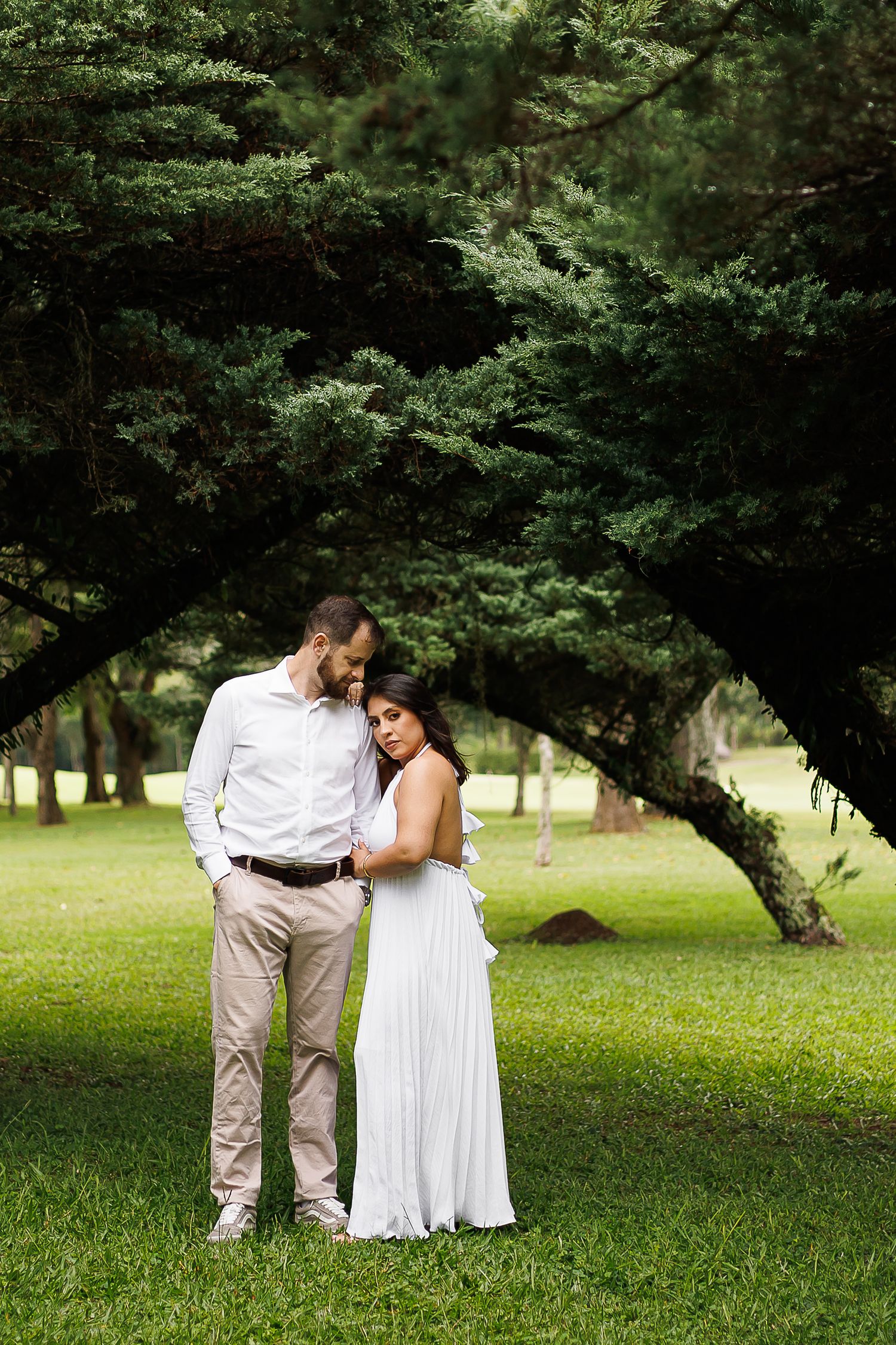Couple in white attire embracing under lush green trees in a park during an outdoor engagement photo session.