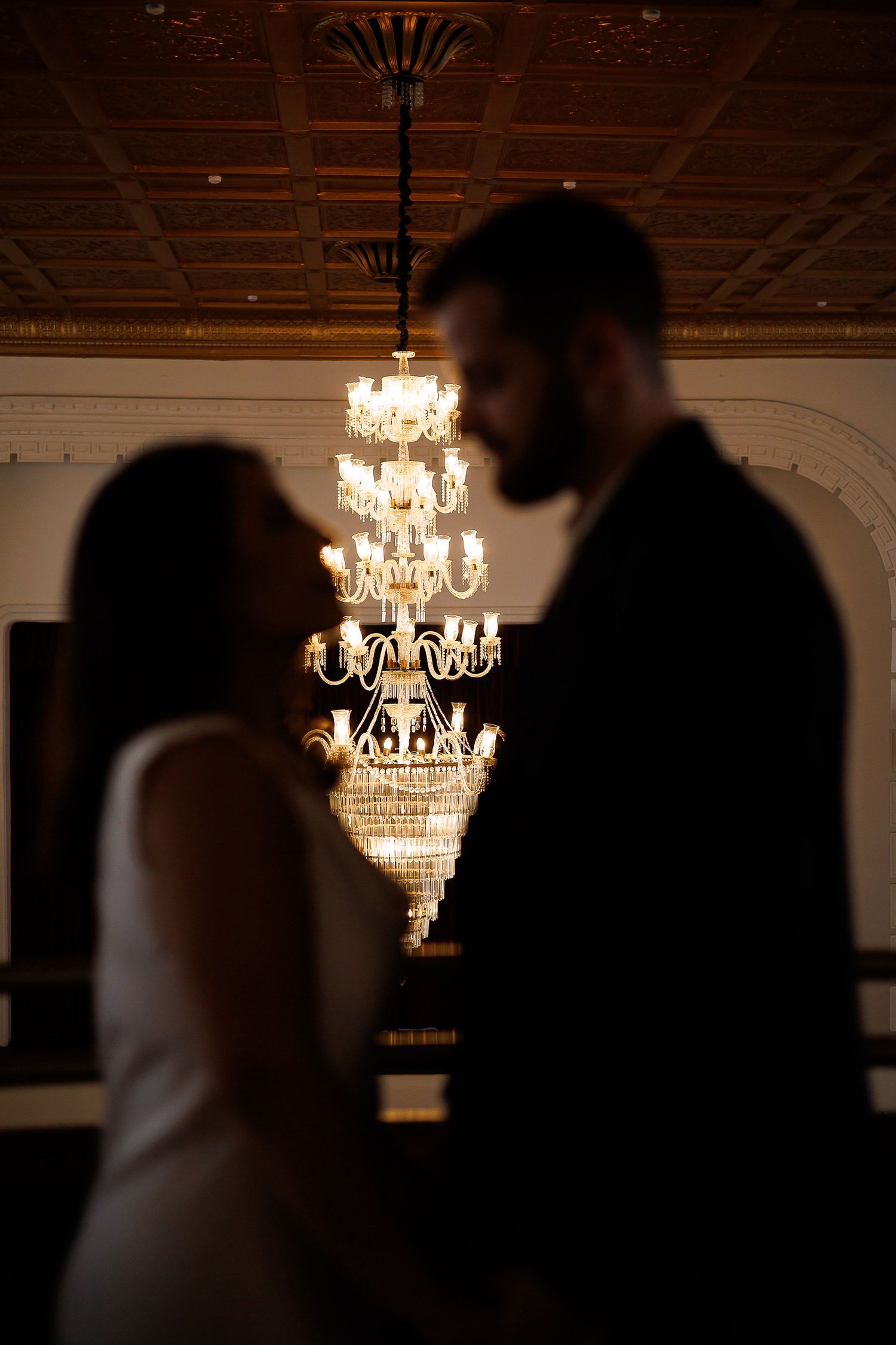 Silhouetted bride and groom facing each other with elegant crystal chandelier glowing between them in ornate ballroom.