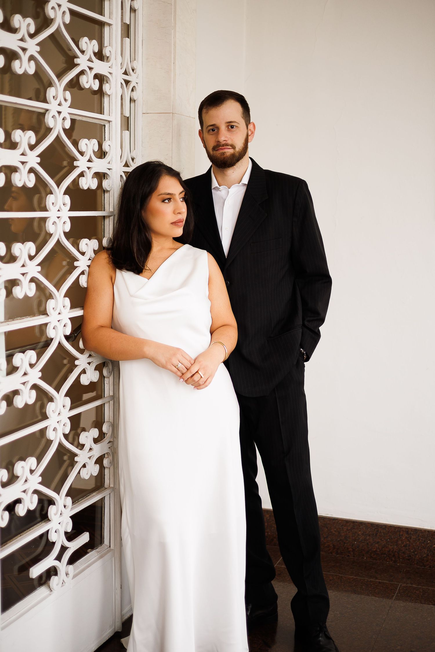 Elegant couple posing by ornate white gate, woman in white gown and man in black suit, formal portrait photography.