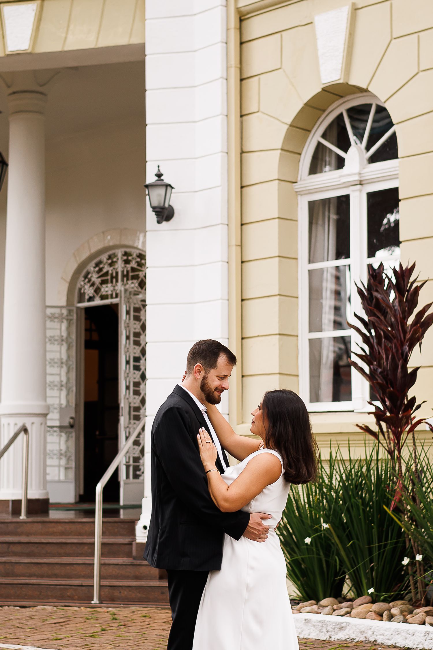 Bride and groom embrace romantically outside an elegant yellow and white building with arched windows and lush greenery.