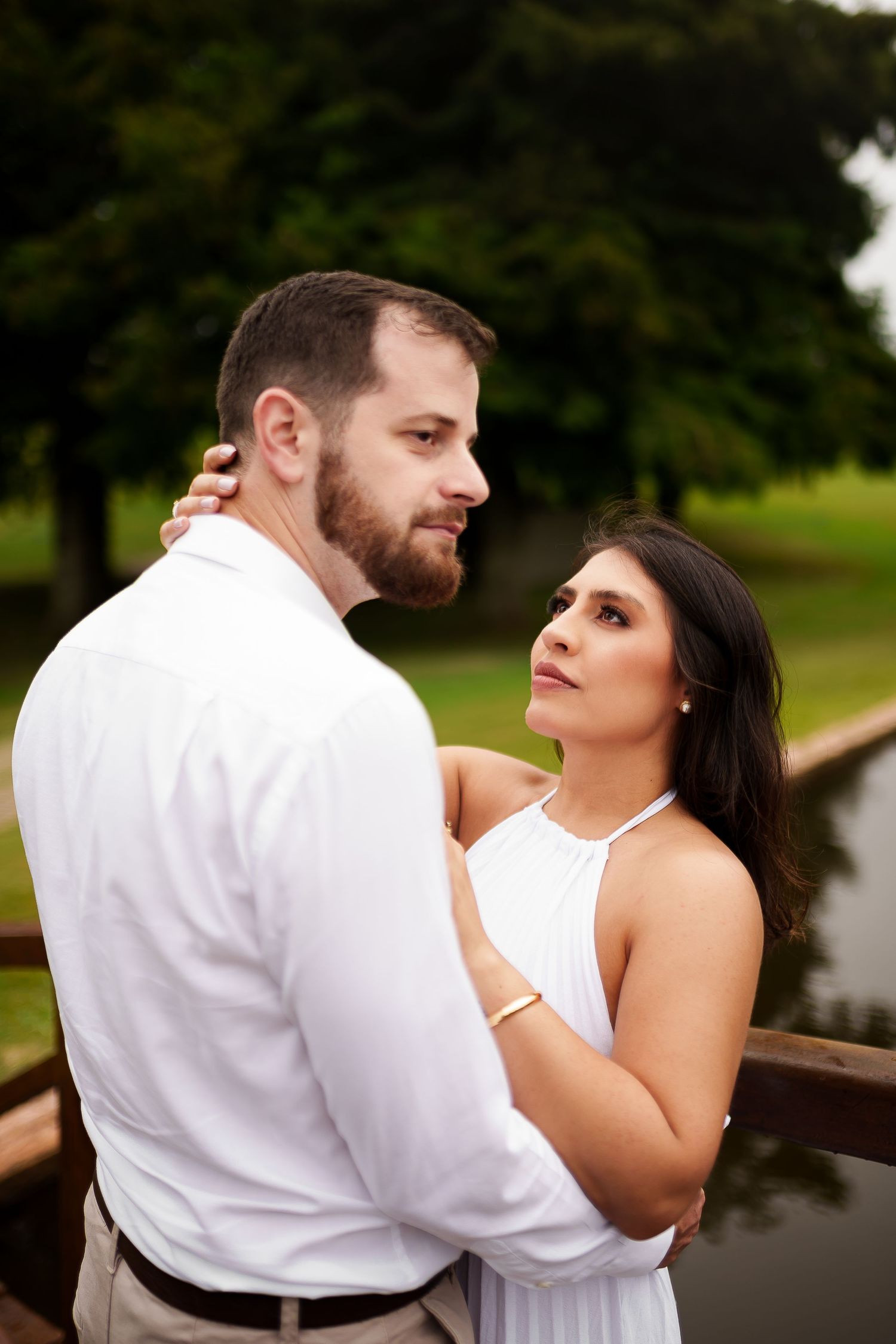 A couple in white attire embraces on a wooden bridge, the woman gazing lovingly at the bearded man outdoors.