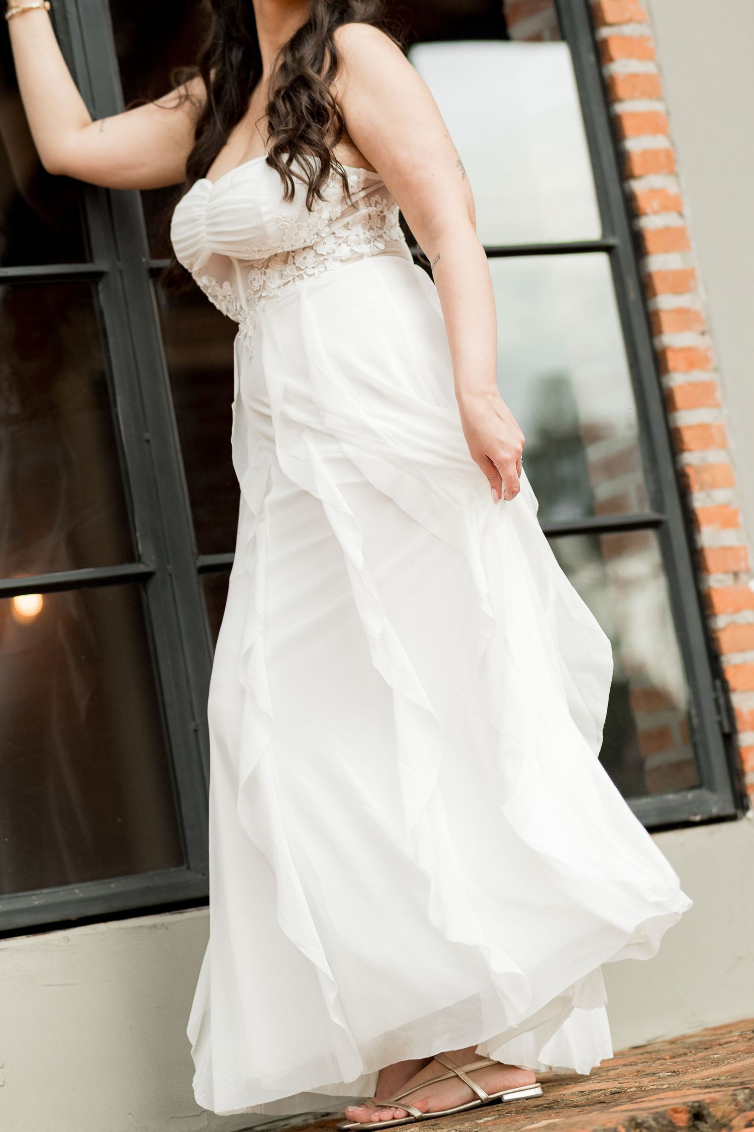 Woman in strapless white wedding gown with lace bodice and flowing tiered skirt posing near brick wall.
