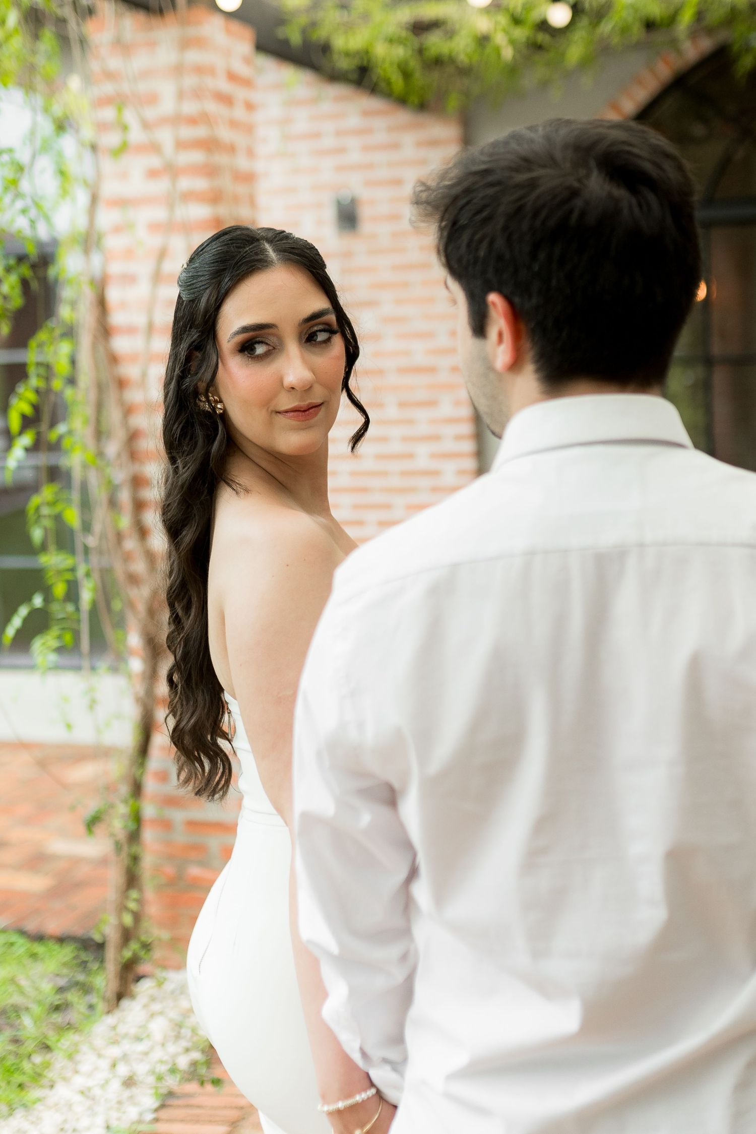 Bride in backless white gown gazes over shoulder at groom in white shirt outdoors near brick wall.