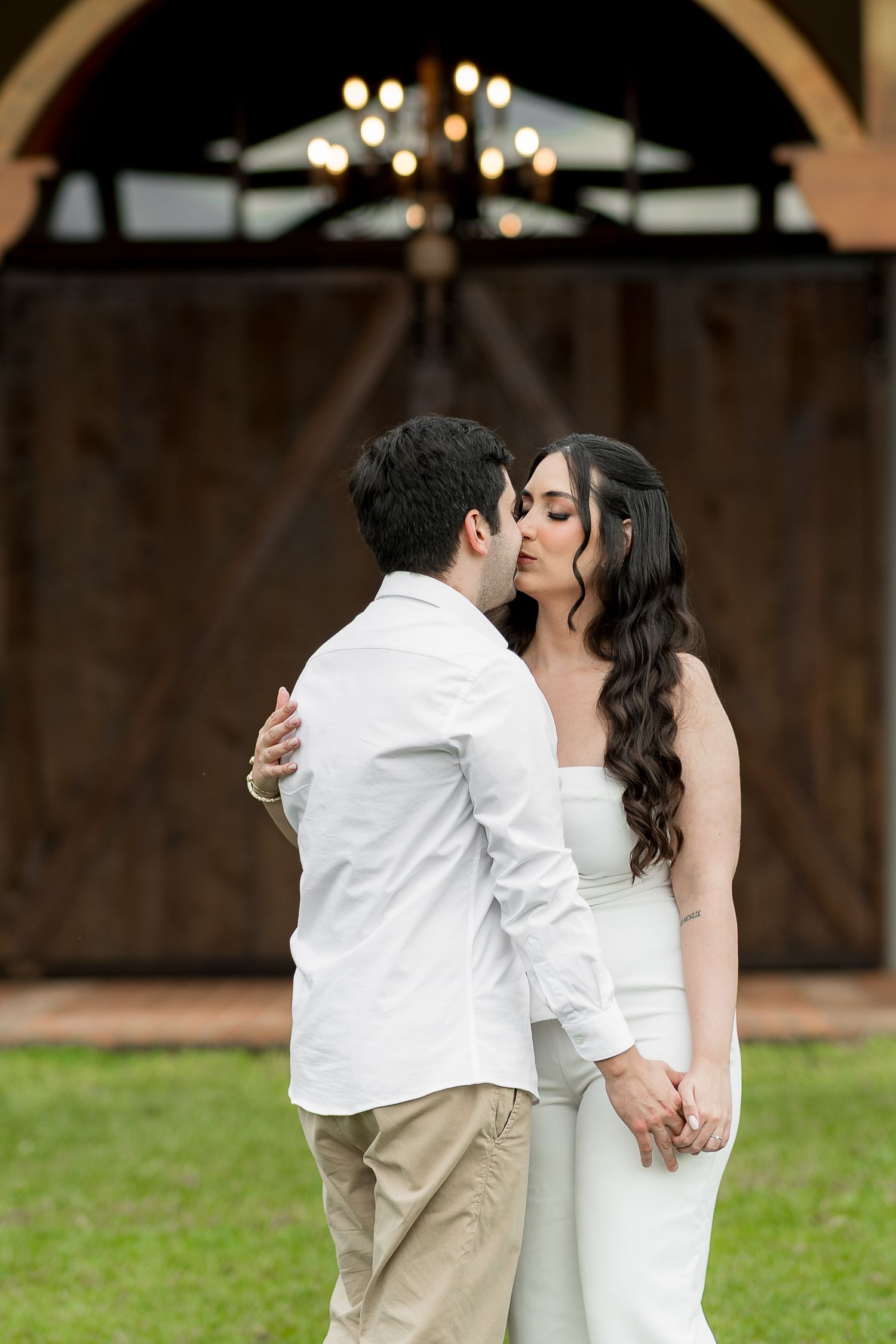 Couple sharing romantic kiss outdoors, man in white shirt and woman in white dress, with barn doors and chandelier behind.