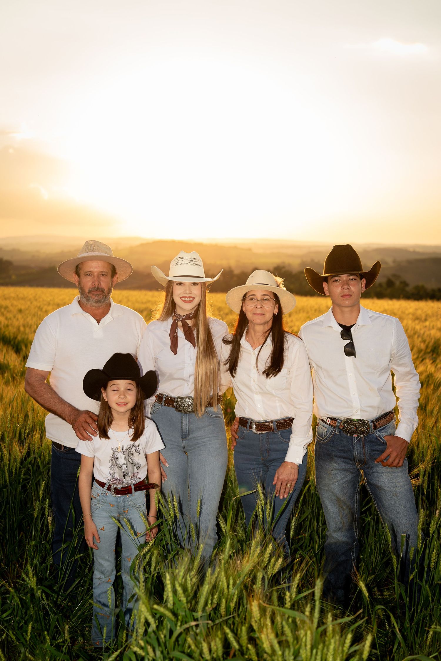 Family of five in cowboy hats posing in a golden wheat field at sunset wearing white shirts and jeans.