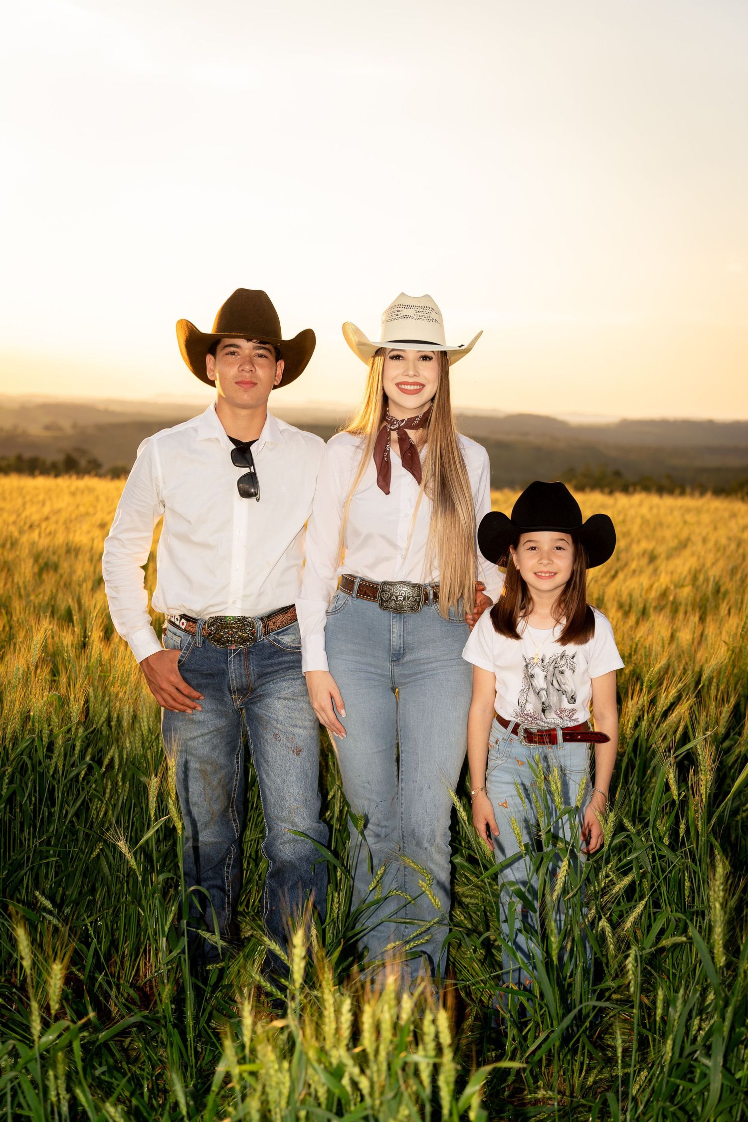 Three people in cowboy hats and jeans pose in a golden wheat field at sunset, wearing western attire.