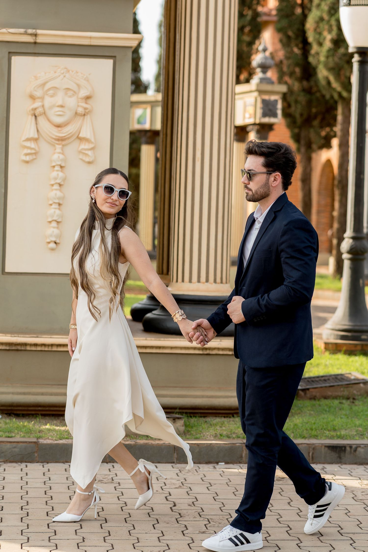 Elegant couple holding hands outdoors, woman in white dress and sunglasses, man in navy suit near ornate stone relief.