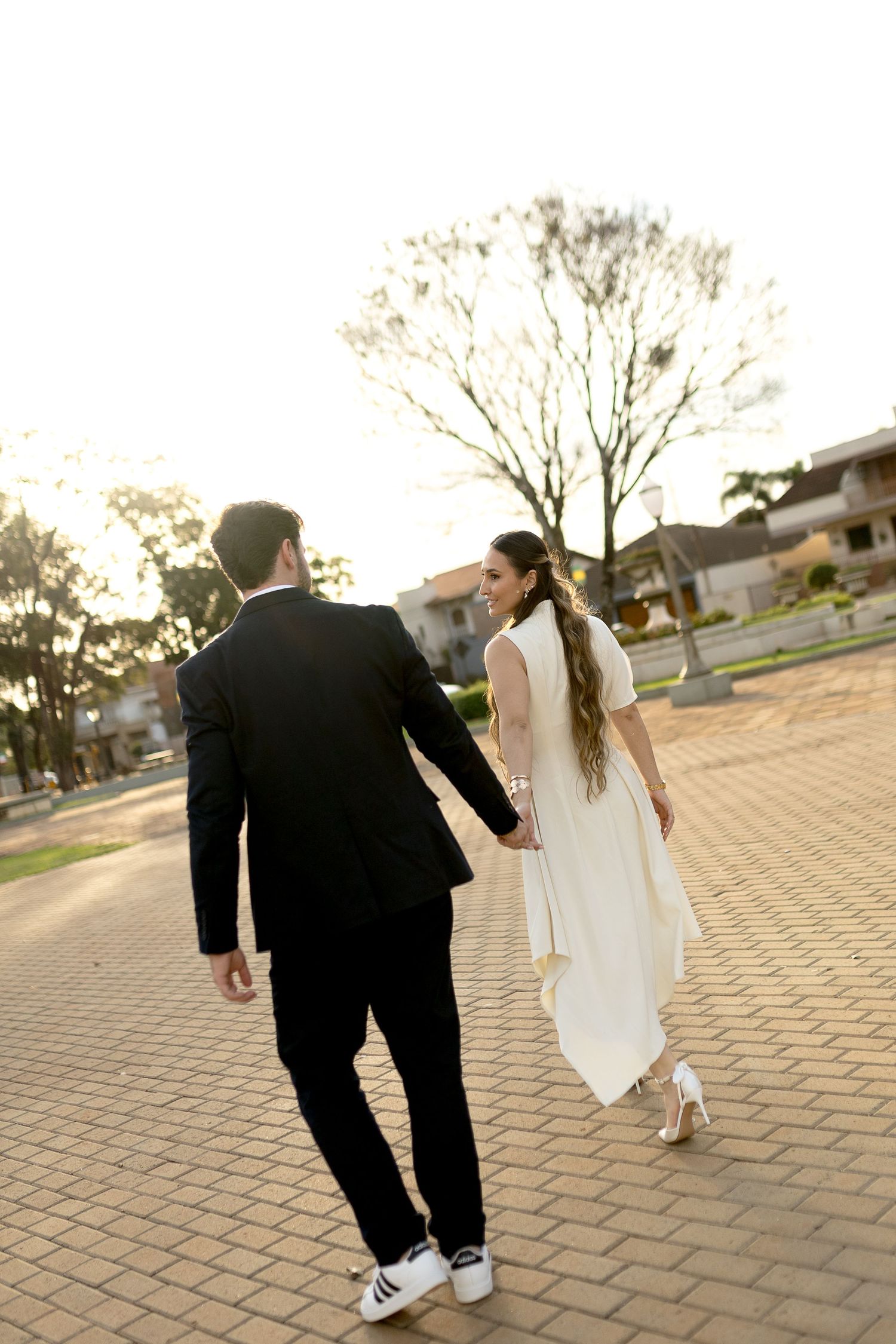 Bride in white dress and groom in black suit holding hands walking on brick path at golden hour.