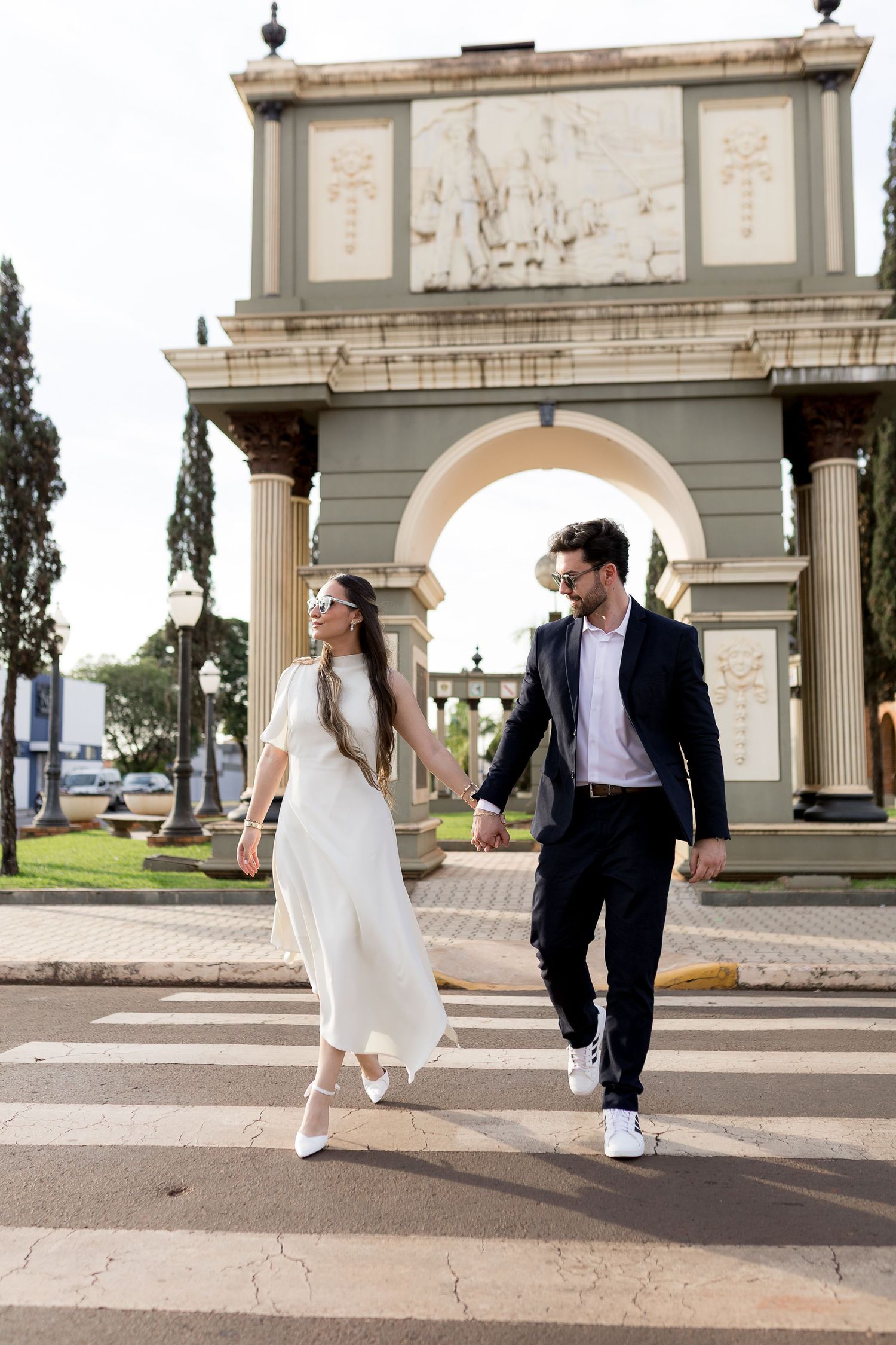 Elegant couple in wedding attire holding hands while crossing a zebra crossing near a classical arch monument.