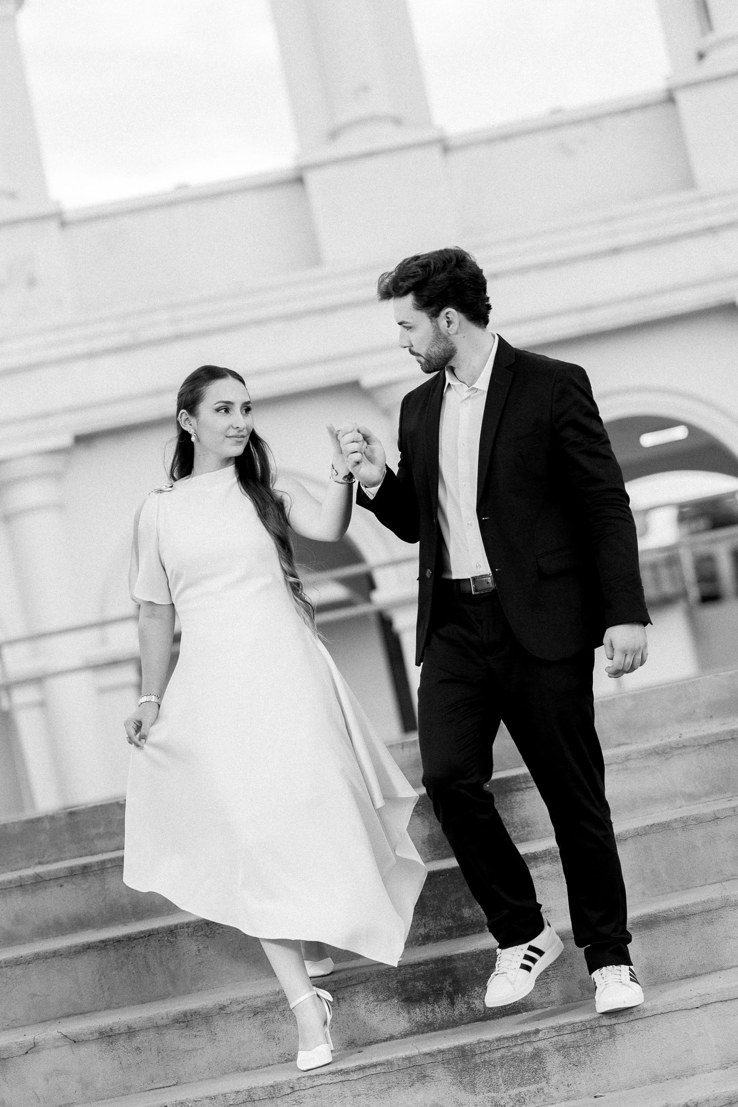Black and white photo of an elegant couple holding hands while descending outdoor steps, woman in white dress, man in dark suit.