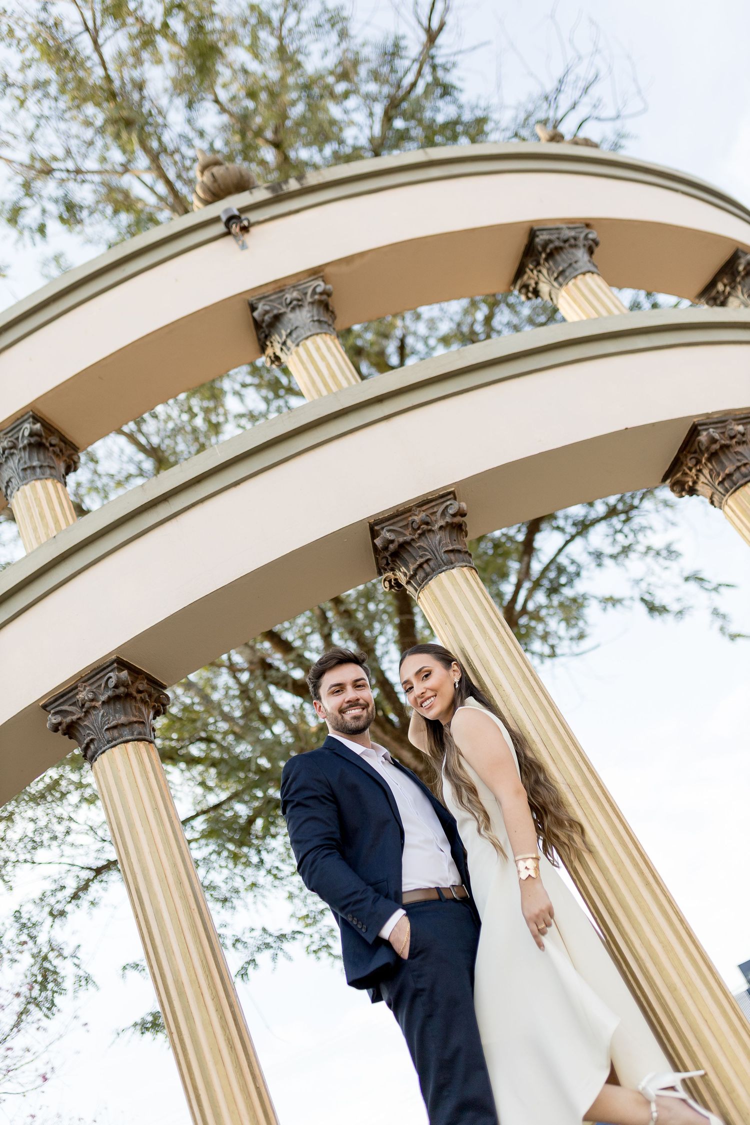 Smiling couple in wedding attire posing under elegant arched columns in a park setting.