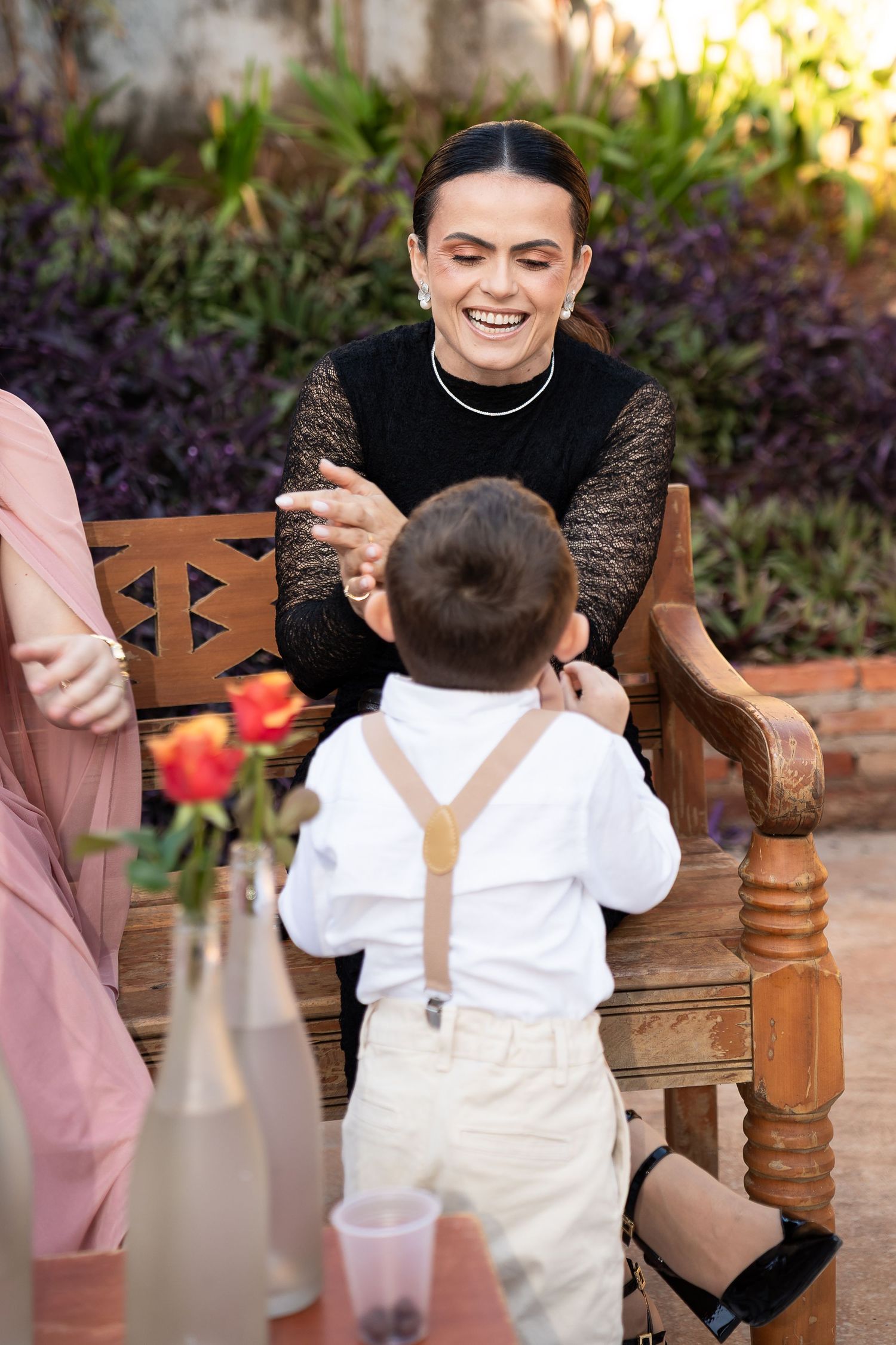 A laughing woman in black lace top interacts with a toddler in suspenders at an outdoor garden party.