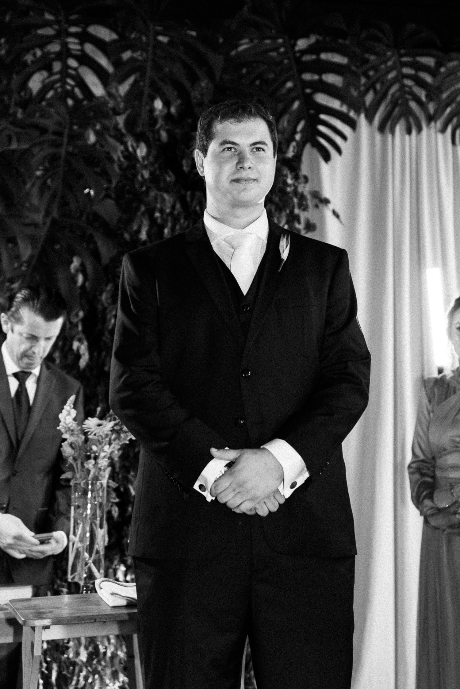 Groom in black suit standing at wedding altar with hands clasped, black and white photo.