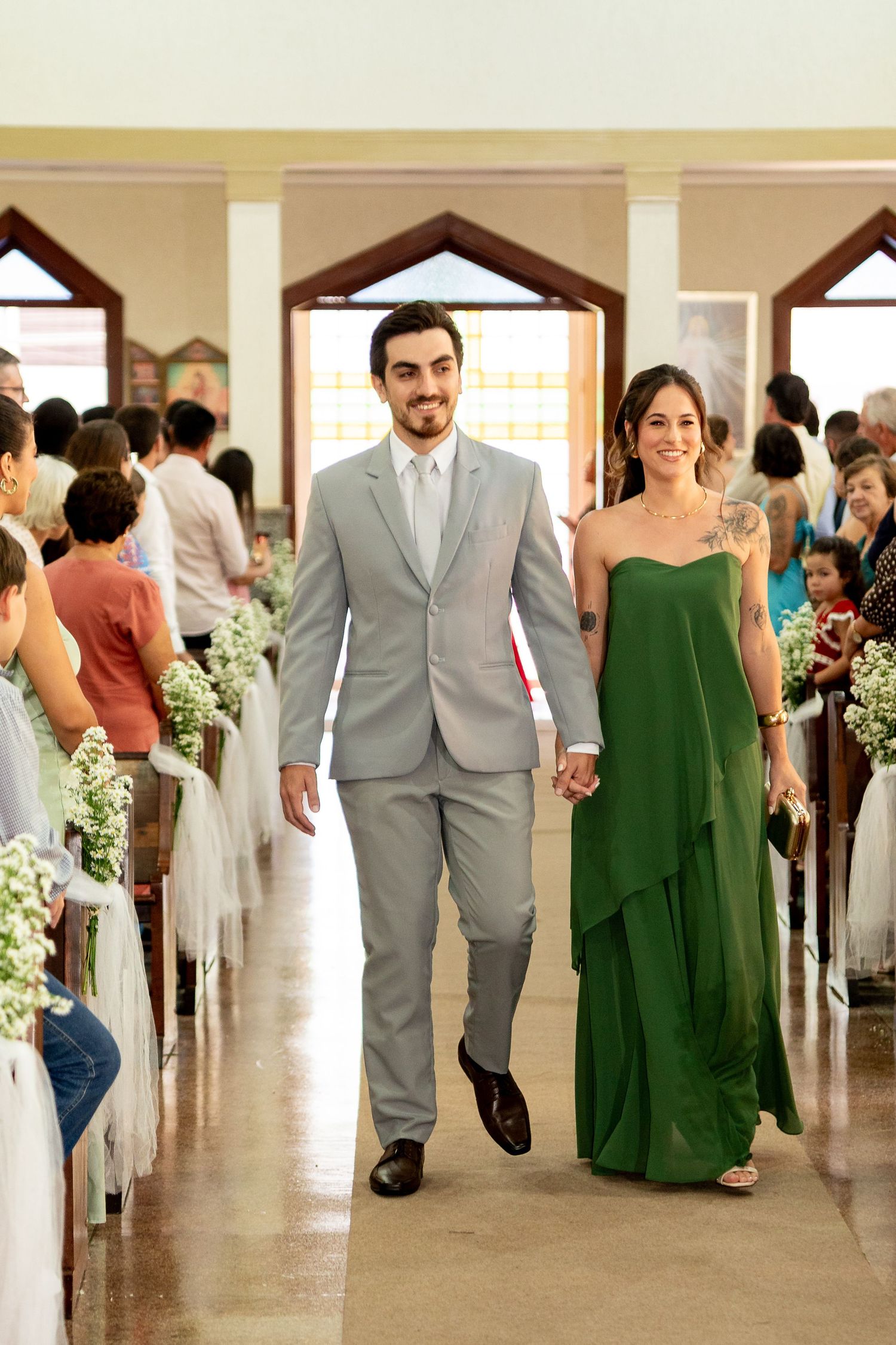 A couple walks hand-in-hand down a church aisle, man in gray suit, woman in green gown, guests seated on both sides.