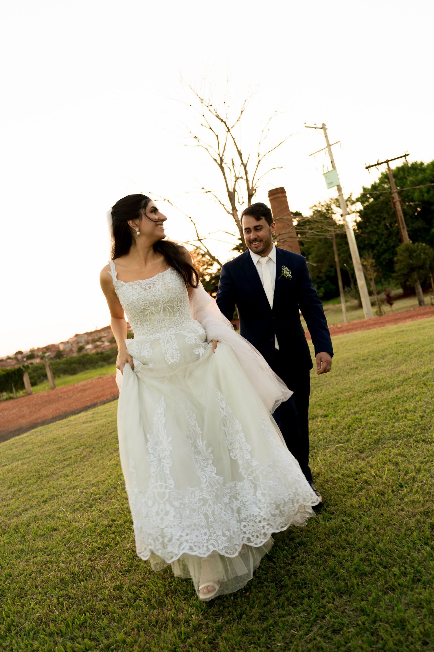 Bride in white lace gown and groom in navy suit walk together on green lawn during golden hour outdoor wedding.