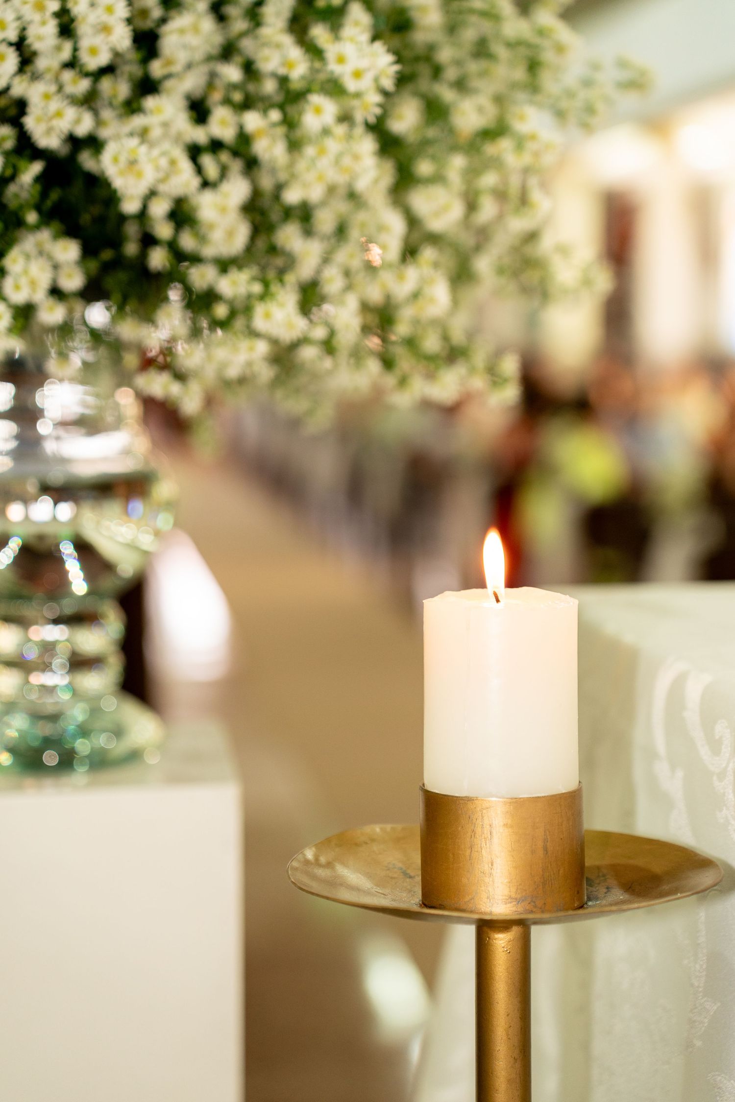 A lit white pillar candle on a gold stand with white floral arrangements in a softly blurred wedding venue background.