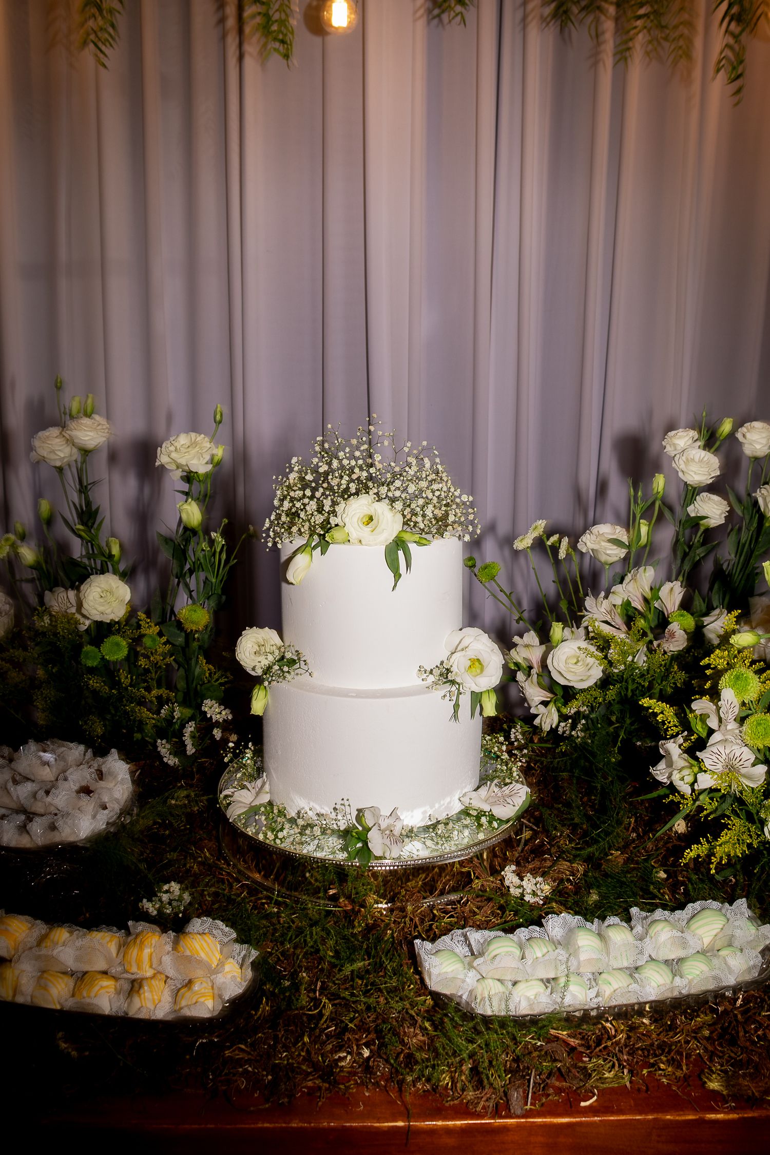 Elegant two-tier white wedding cake adorned with white flowers on a dessert table with floral arrangements.