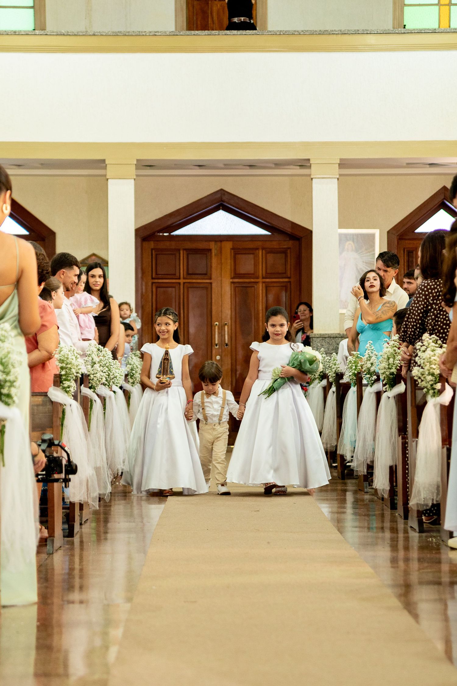 Children in white dresses process down church aisle at wedding ceremony.