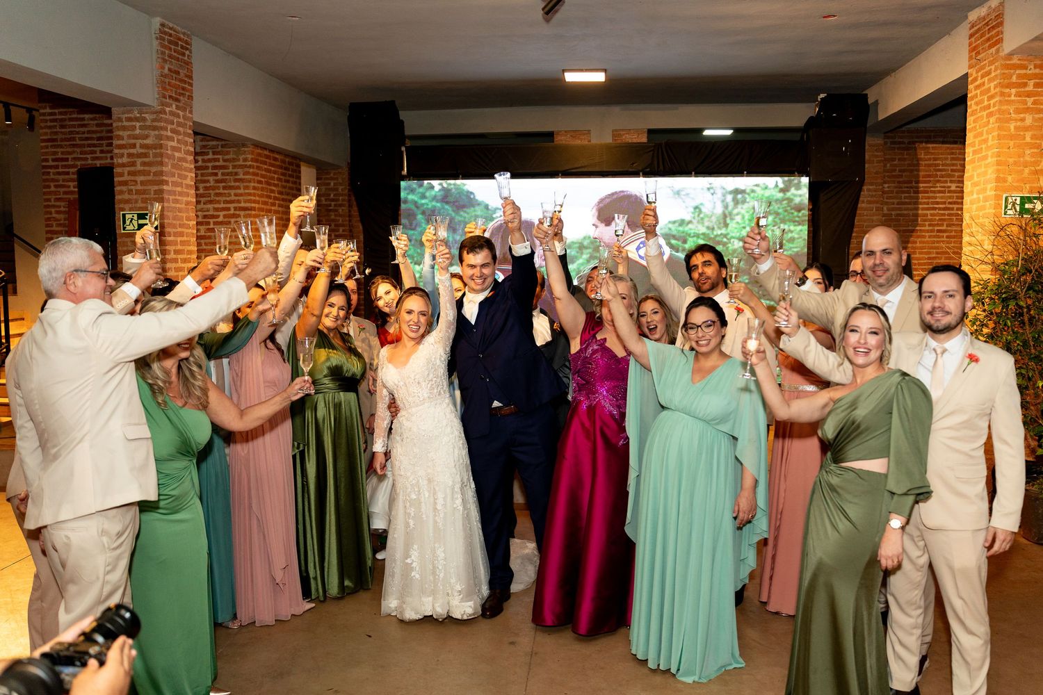 Wedding party celebrating with raised hands in elegant venue, bride in white gown surrounded by bridesmaids in green and pink dresses.