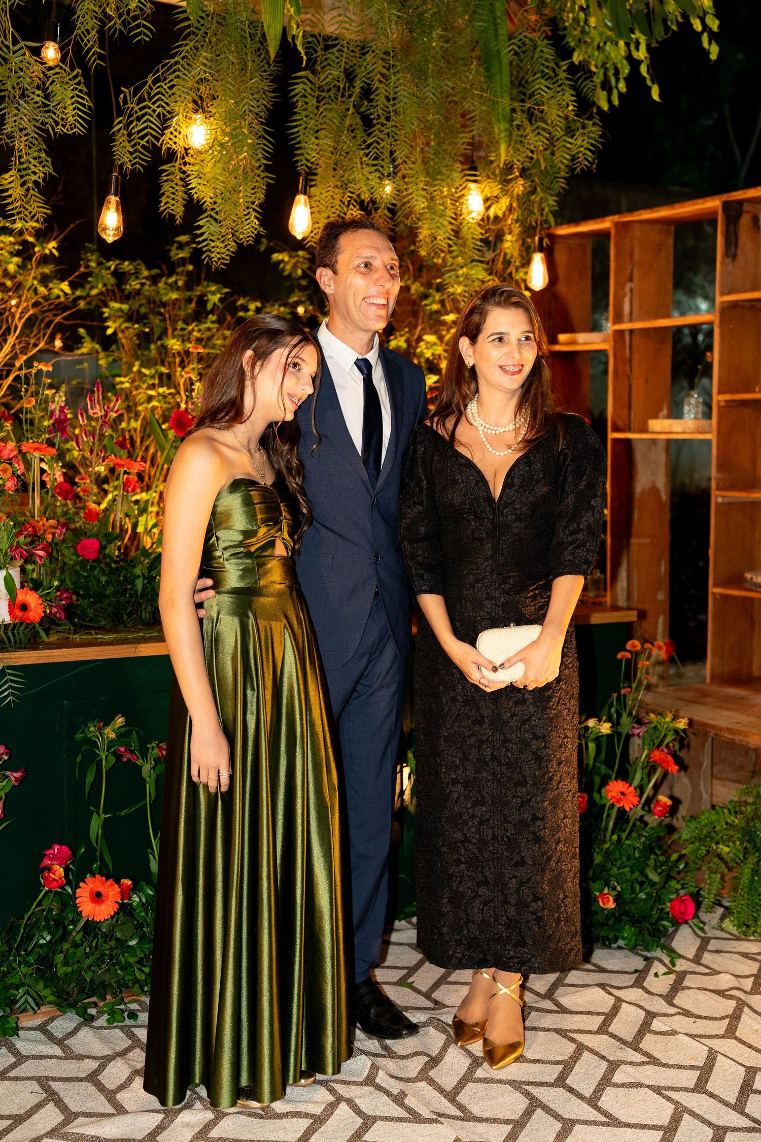 Three elegantly dressed guests pose together at a formal event, surrounded by warm string lights and tropical greenery.