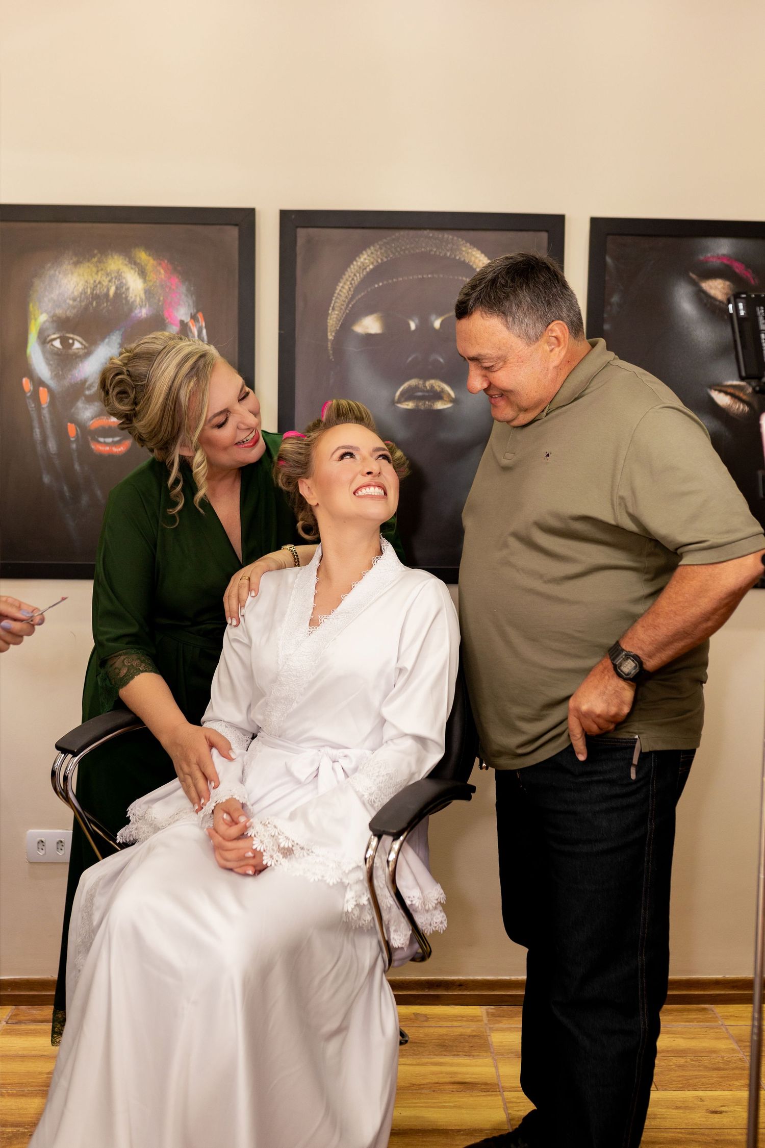 Bride in white robe smiles while getting ready, surrounded by two people in a photo studio.