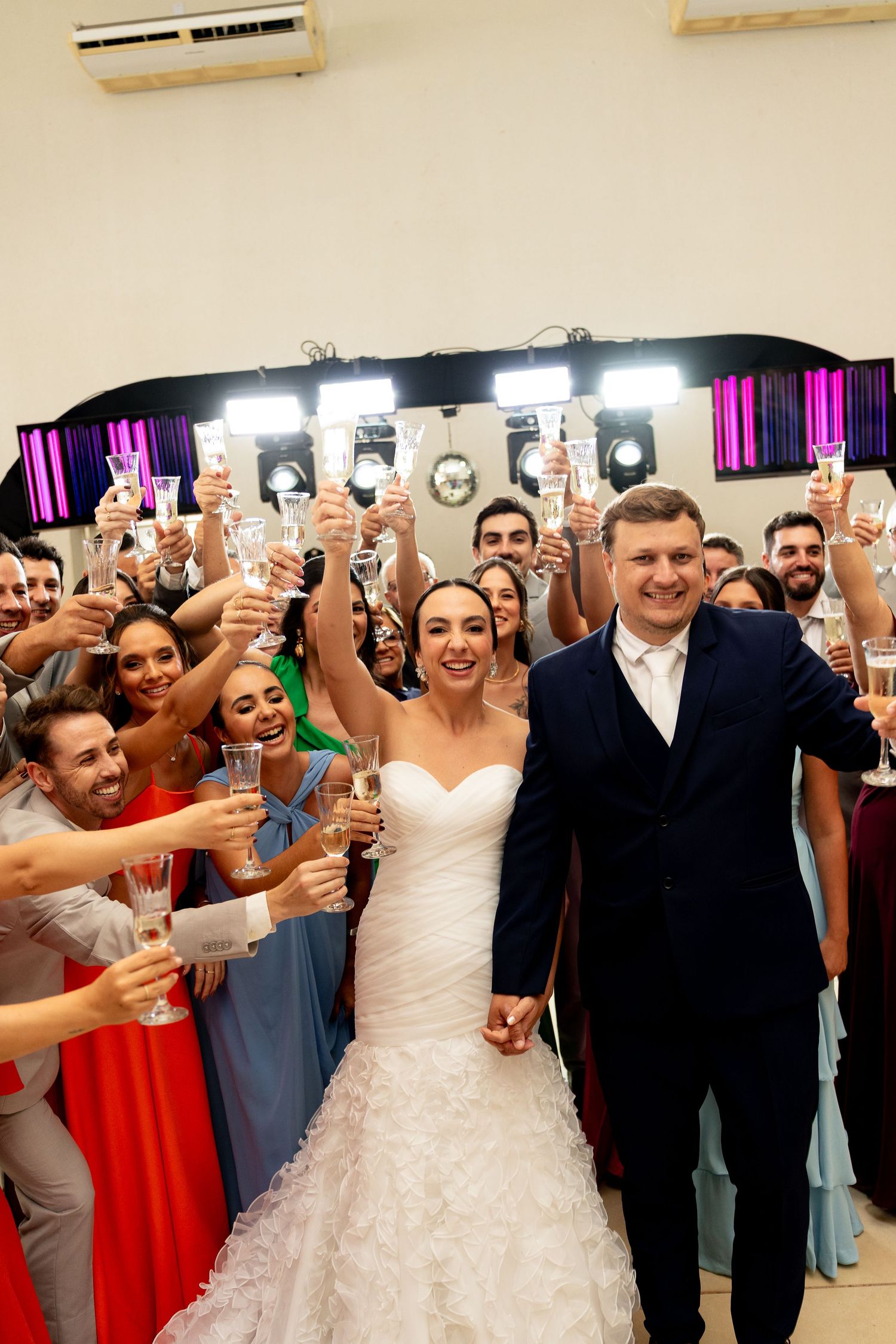 Bride and groom holding champagne glasses celebrate with wedding guests in colorful attire at a reception venue.
