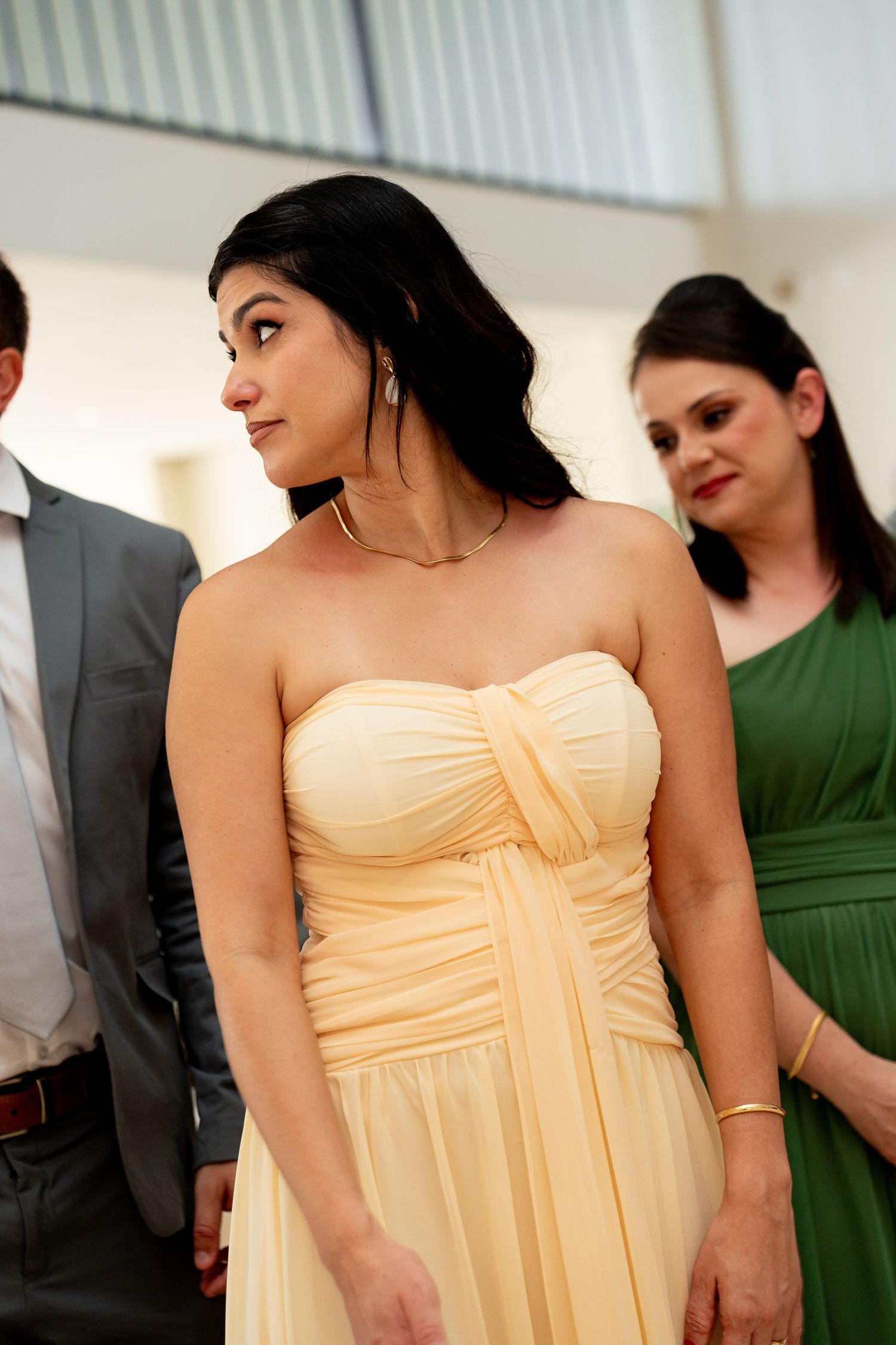 Woman in yellow strapless gown at formal event, with guests in green dress and gray suit behind her.
