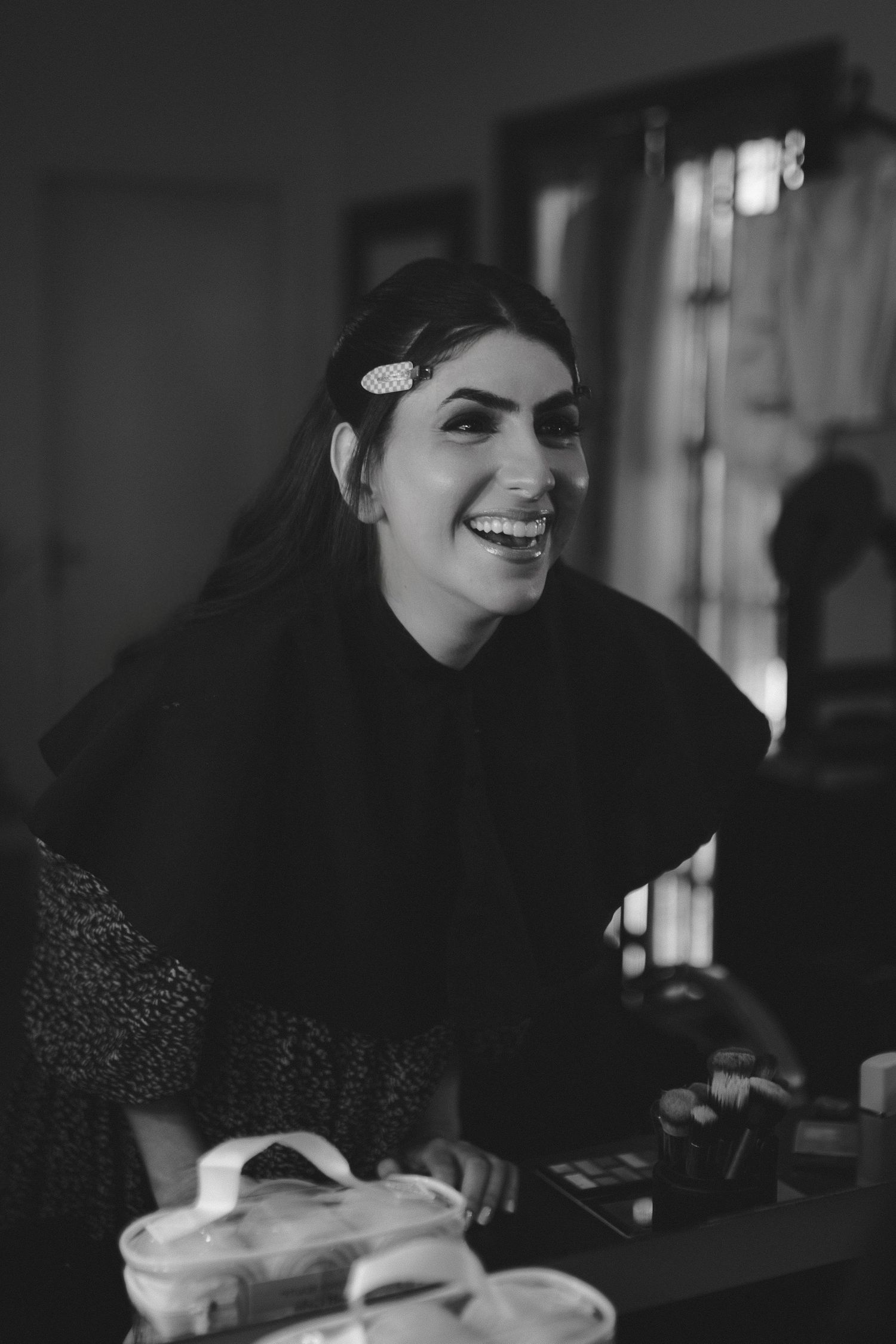 Black and white photo of a laughing woman wearing a hijab and hair clip, standing near a makeup table indoors.
